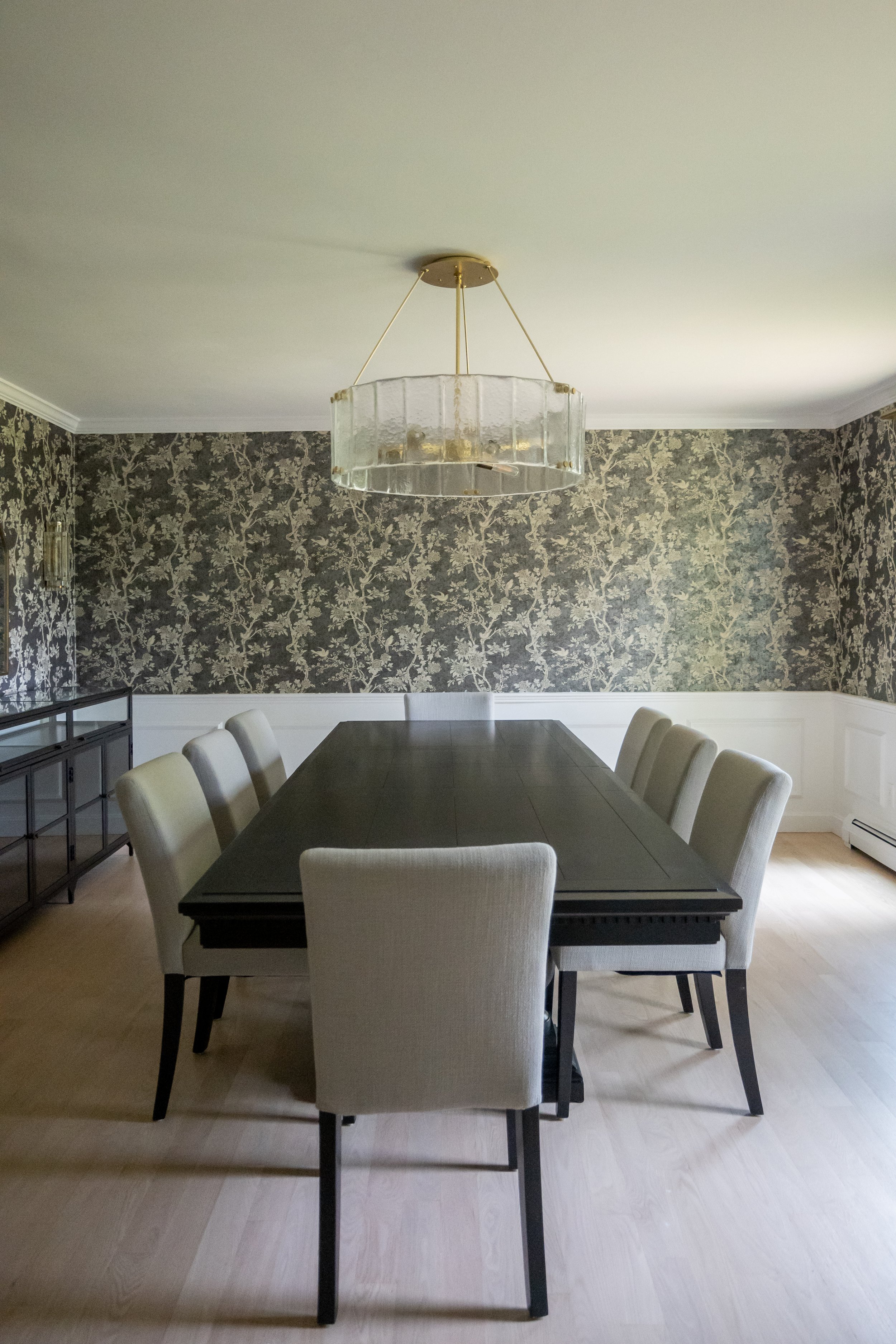 Large dark wood dining table surrounded by eight upholstered chairs, showcased beneath a large circular glass chandelier and green floral wallpaper.