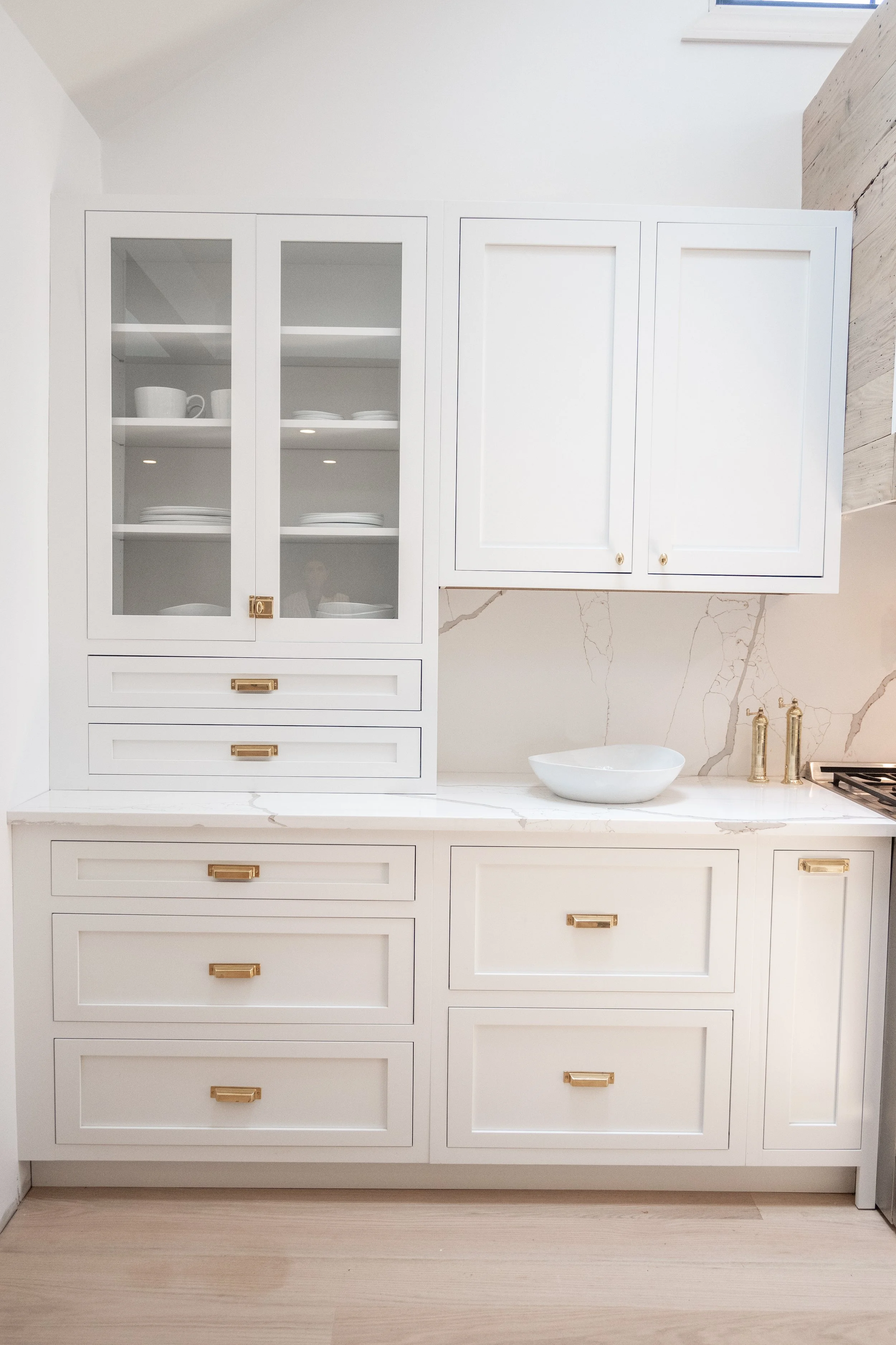 Close-up of luxury white kitchen cabinets styled with gold knobs, light marble countertops, and a matching marble backsplash.