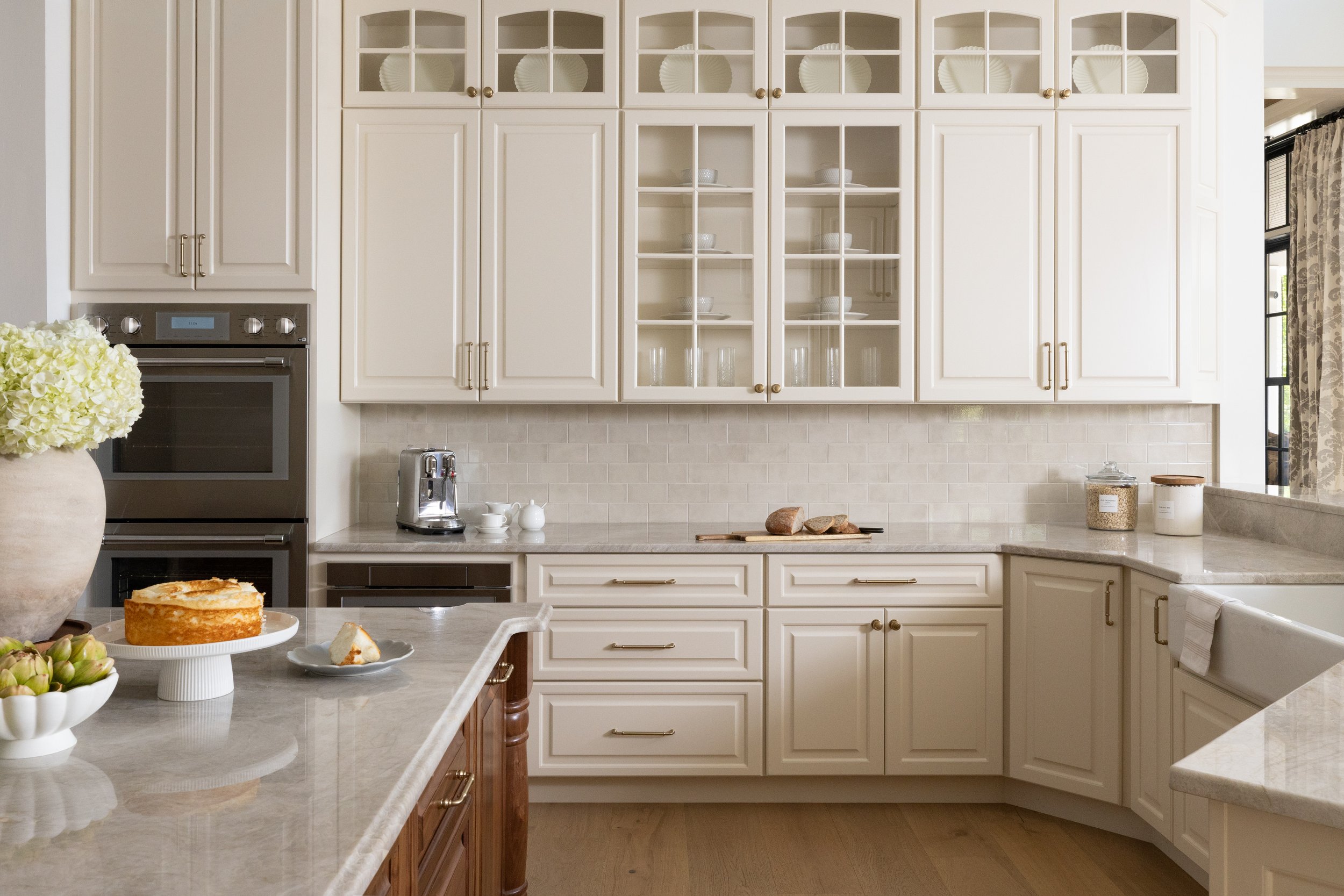 The image shows a modern kitchen with white cabinets and a beige countertop. There are glass-front upper cabinets displaying white dishes and bowls. On the counter, there is a silver coffee machine, a small white jar, and a glass container. The kitch