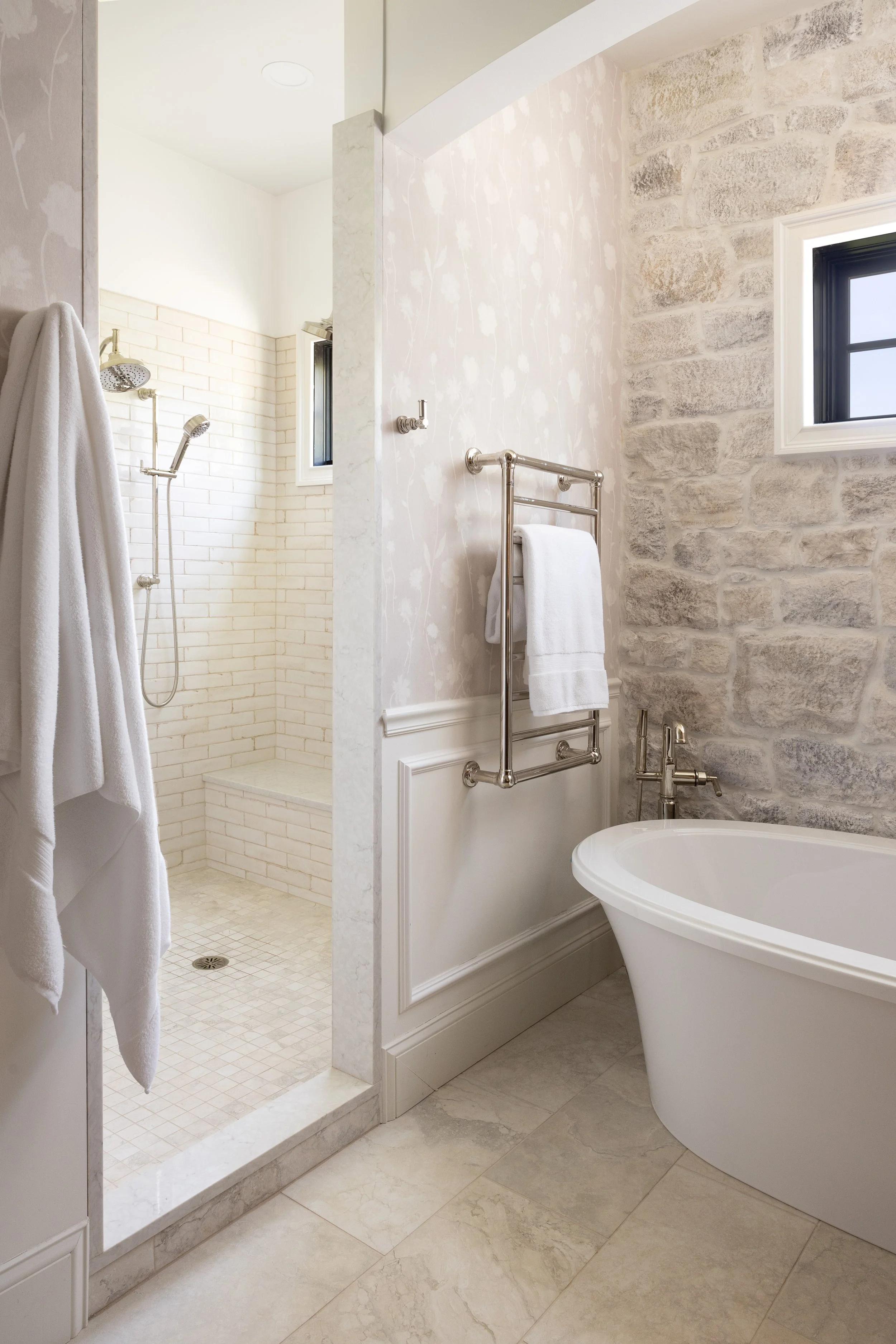 A bathroom with a stand-up shower area to the left, with white brick tile walls and a shower head, next to a marble threshold. On the right, there is a white clawfoot bathtub, a towel on a chrome towel rack, and a window with black trim on a stone ac