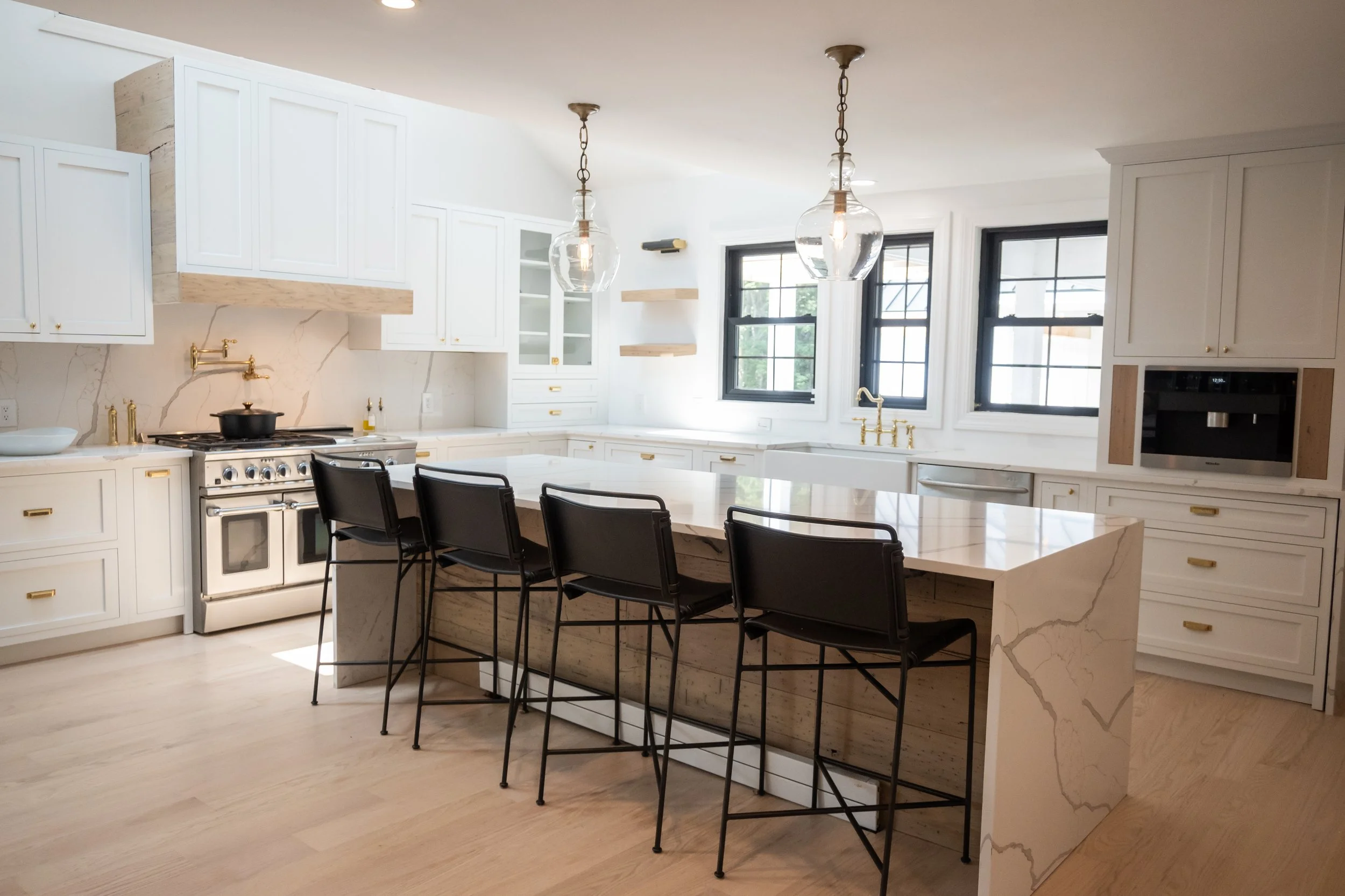 Modern kitchen with white cabinets, a marble island, black chairs at the island, large windows, pendant lights, and gold fixtures.