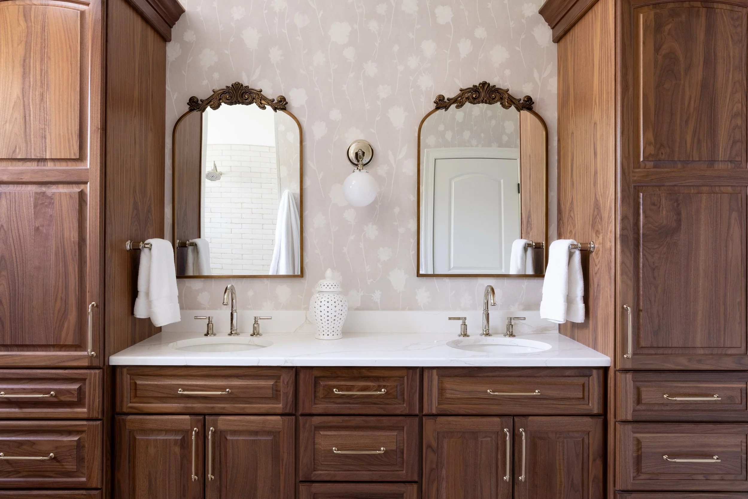 A bathroom vanity with dark wood cabinets, two mirrors with ornate gold frames, a white marble countertop with two sinks, and a white decorative vase in the center. The background wall has wallpaper with a floral pattern, and there are white towels h