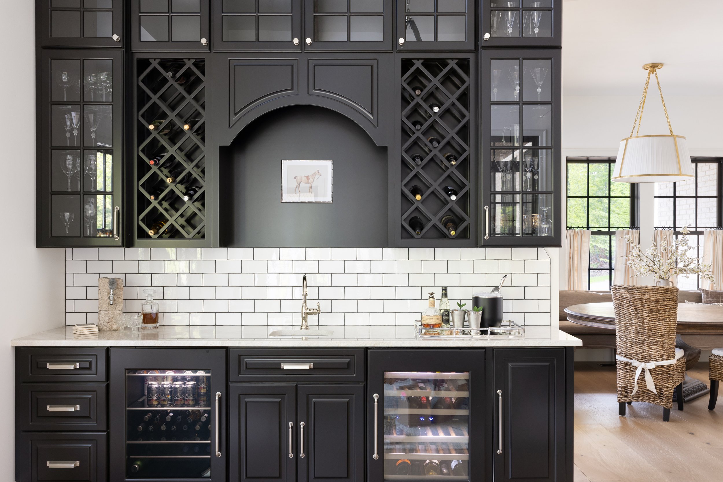 A modern black kitchen cabinet with glass doors and wine racks, set above a white tiled backsplash and countertop, with a small framed horse picture on the wall.