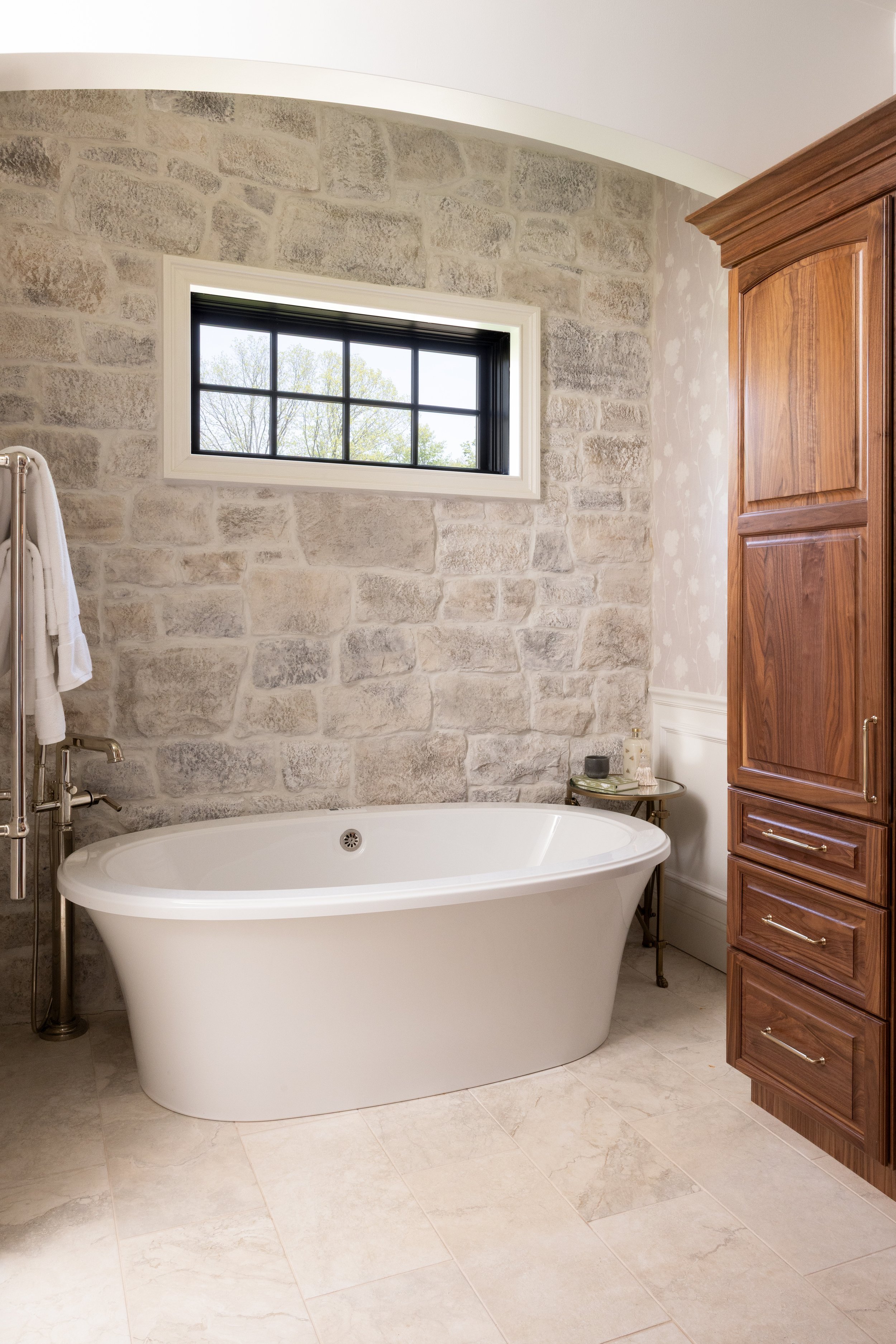 A bathroom with a freestanding bathtub, a window with black grid framing, a stone wall behind the tub, beige floor tiles, a wooden cabinet on the right, and a small side table with toiletries.