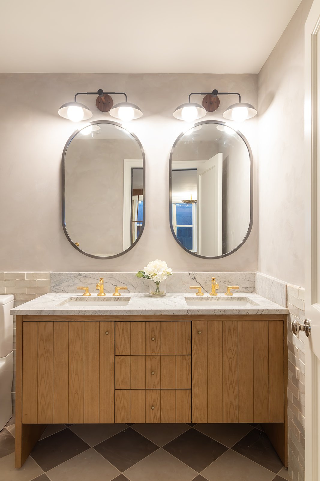 Modern master bathroom featuring his and hers sinks, light marble countertops, warm brown cabinets, golden faucets, and twin vertically oval wall-mounted mirrors.