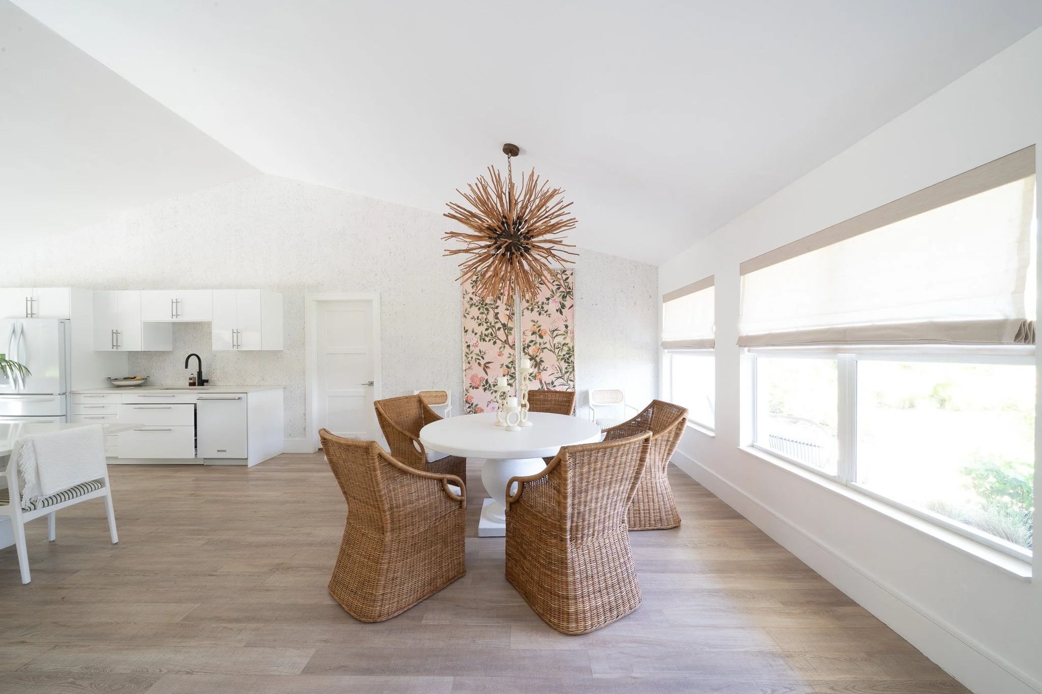 Bright dining space featuring a round white stone table, woven straw-like chairs, a unique branch-ball light fixture, and a minimalist white kitchen with a matte black faucet.