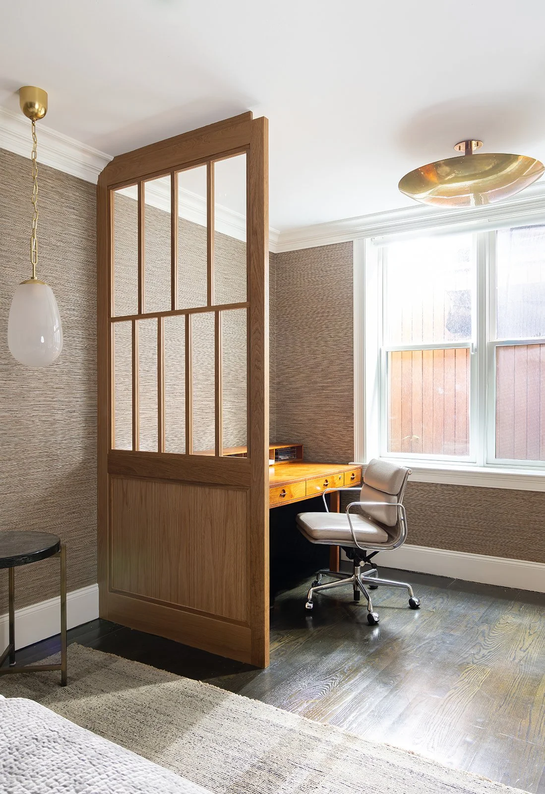 Home office section of a loft bedroom featuring a vintage wooden desk by a bright window, a gray desk chair, dark brown wood flooring, and a gold glass ceiling light.