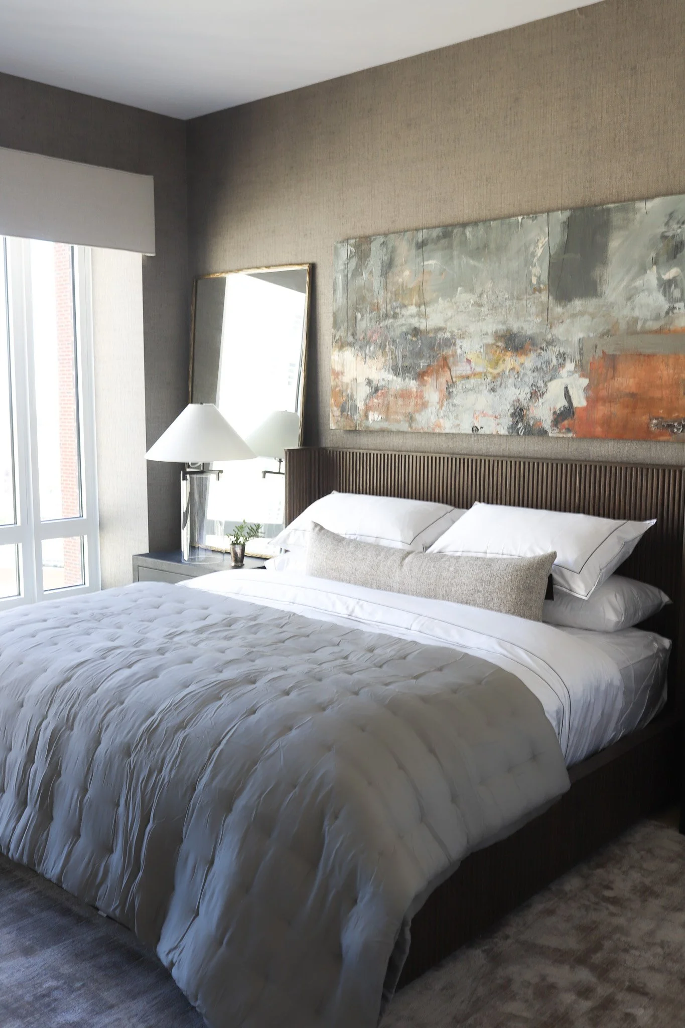 Bright luxury bedroom design with dark beige textured wallpaper, a wooden slatted headboard, contemporary art, and a mirror styled on a bedside table