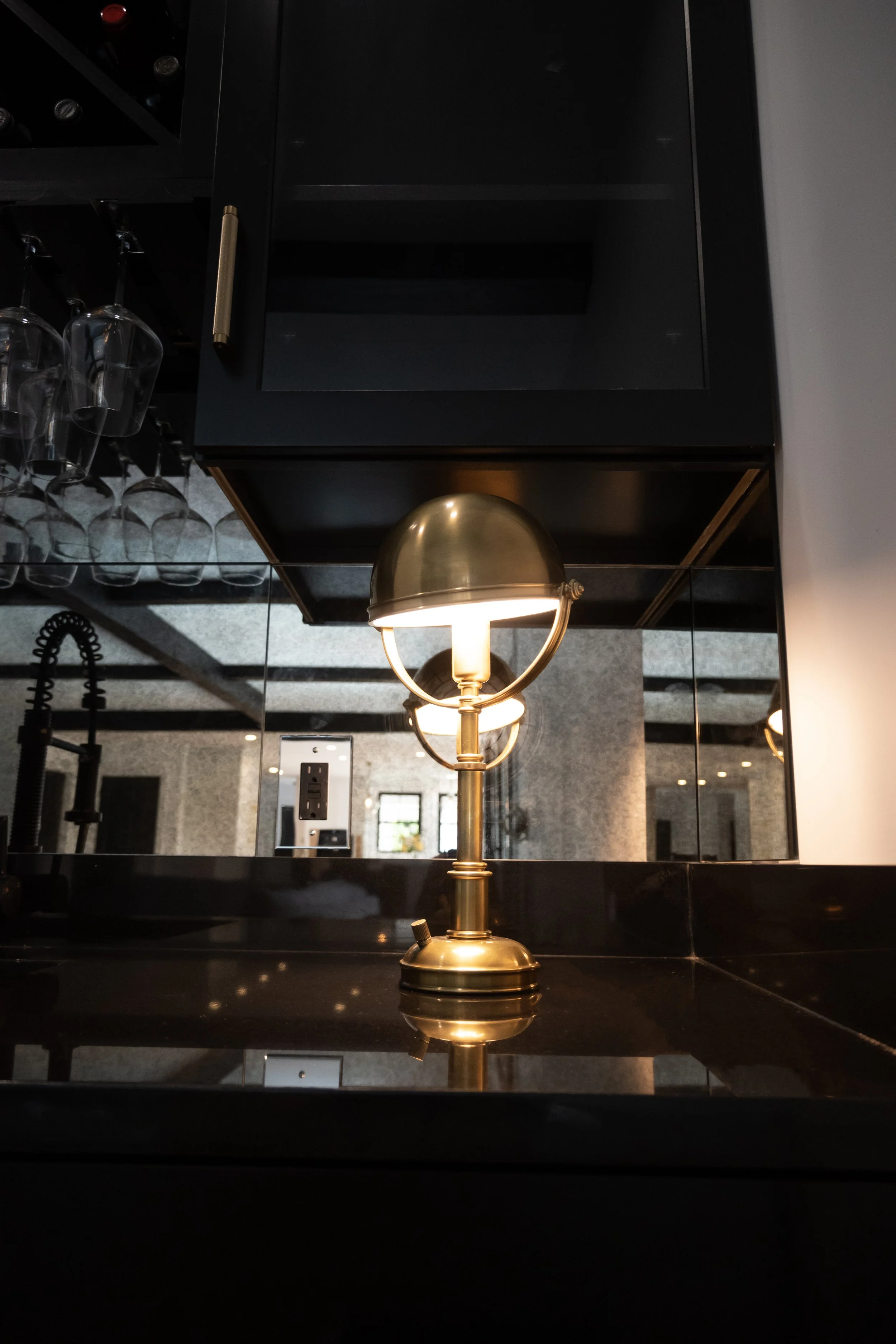 Dark luxury home bar featuring a glowing gold lamp, hanging glassware, an industrial faucet, and a large mirror reflecting textured beige wallpaper.