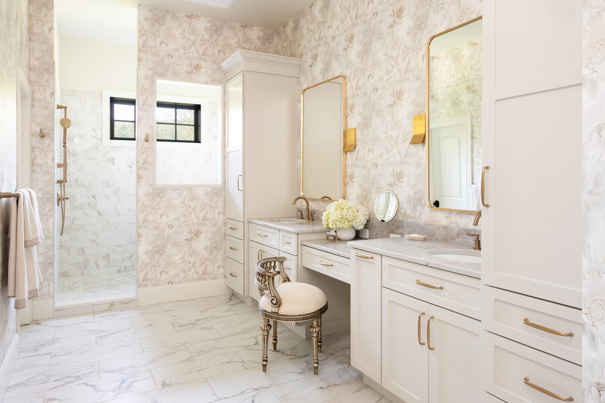 A bright and elegant bathroom with marble flooring and walls, featuring two black-framed windows, a white vanity with gold hardware, a large mirror, a small round mirror, and a vase of white flowers on the countertop.