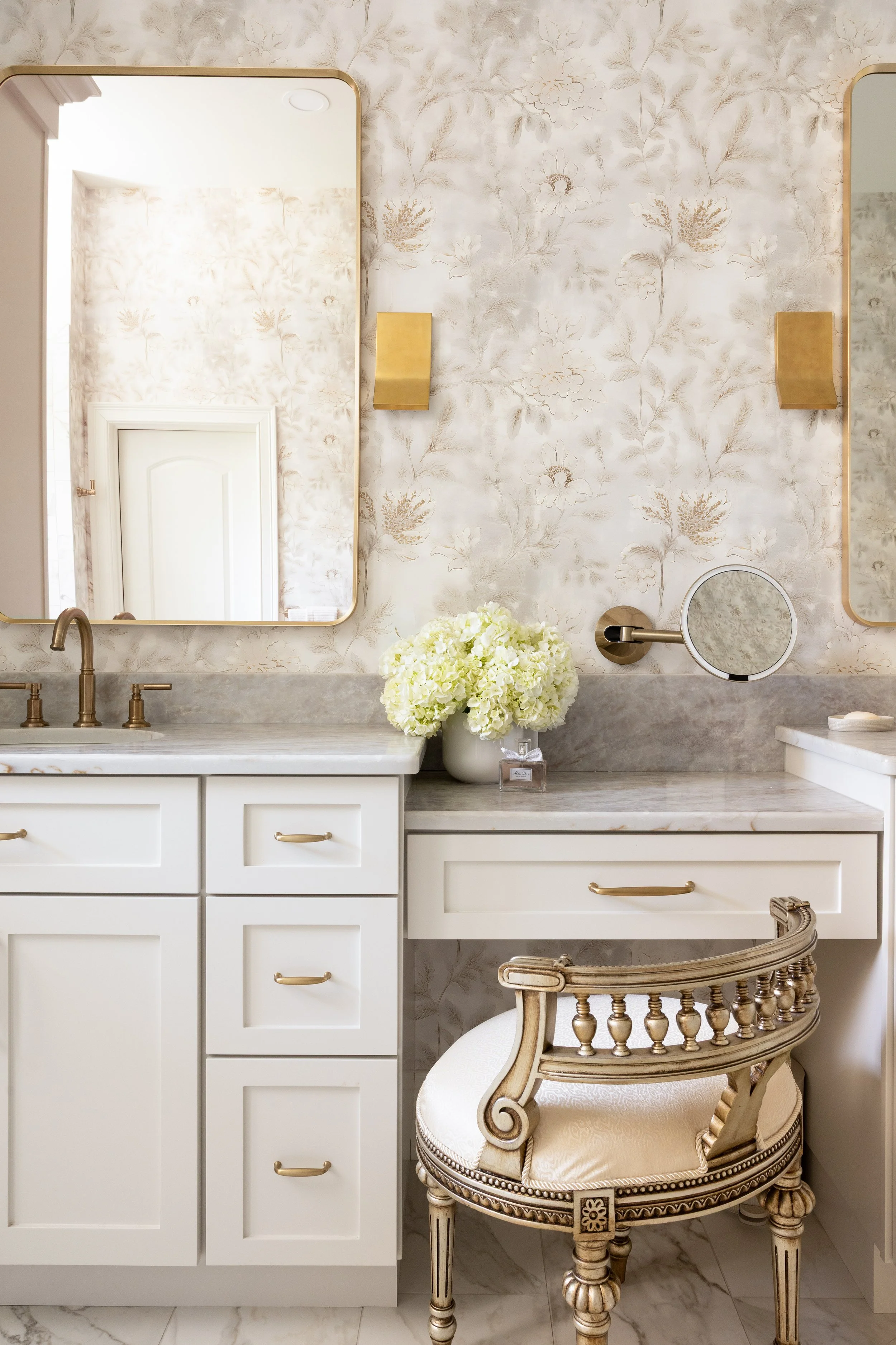 A bathroom vanity with two large mirrors, a white cabinet with gold handles, a marble countertop, a vase of white hydrangeas, a gold wall-mounted makeup mirror, a gold wall sconce, and an ornate vintage-style cream-colored chair with gold accents.