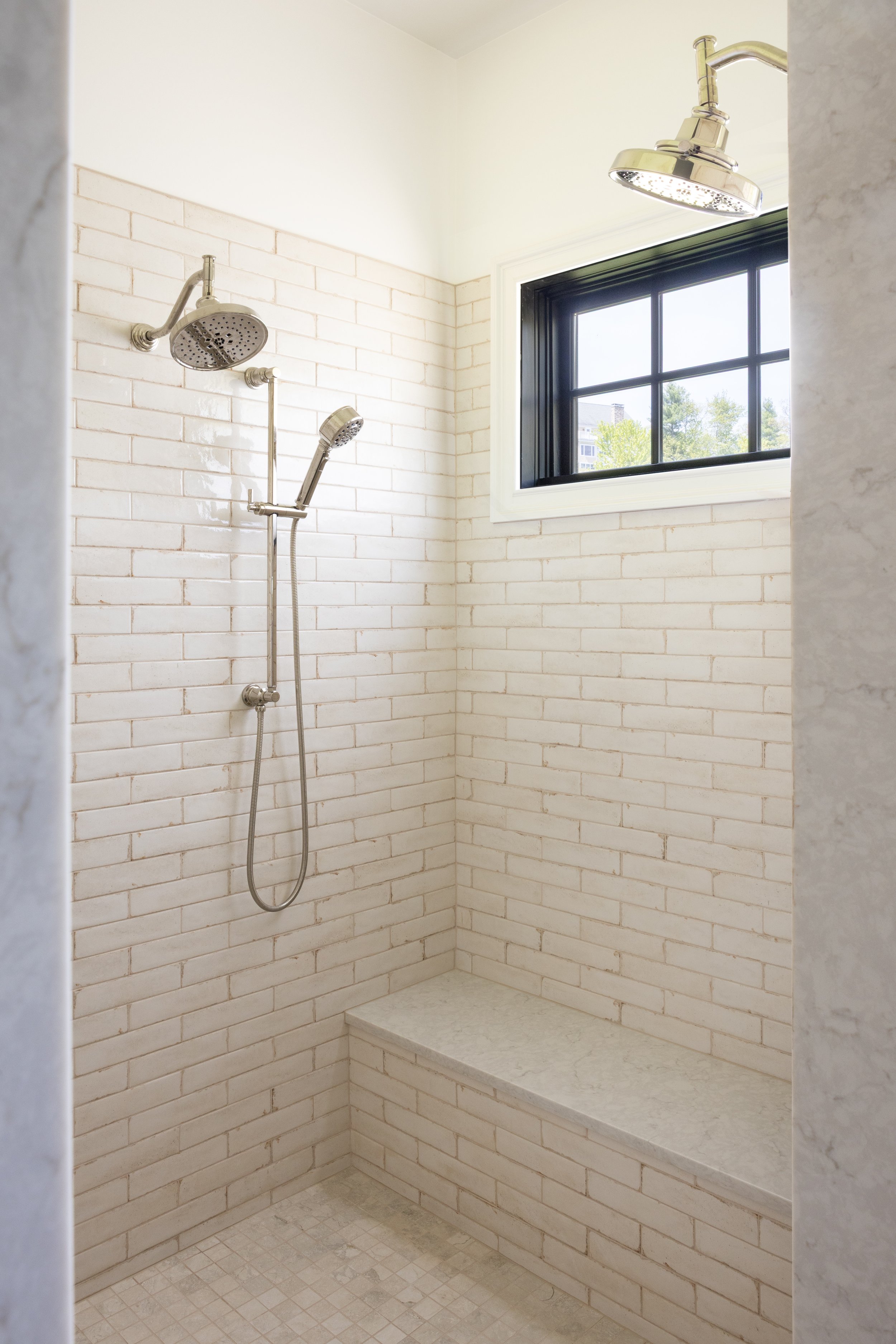 Shower area with white brick walls, two shower heads, a window with black frame, and a built-in marble seat.