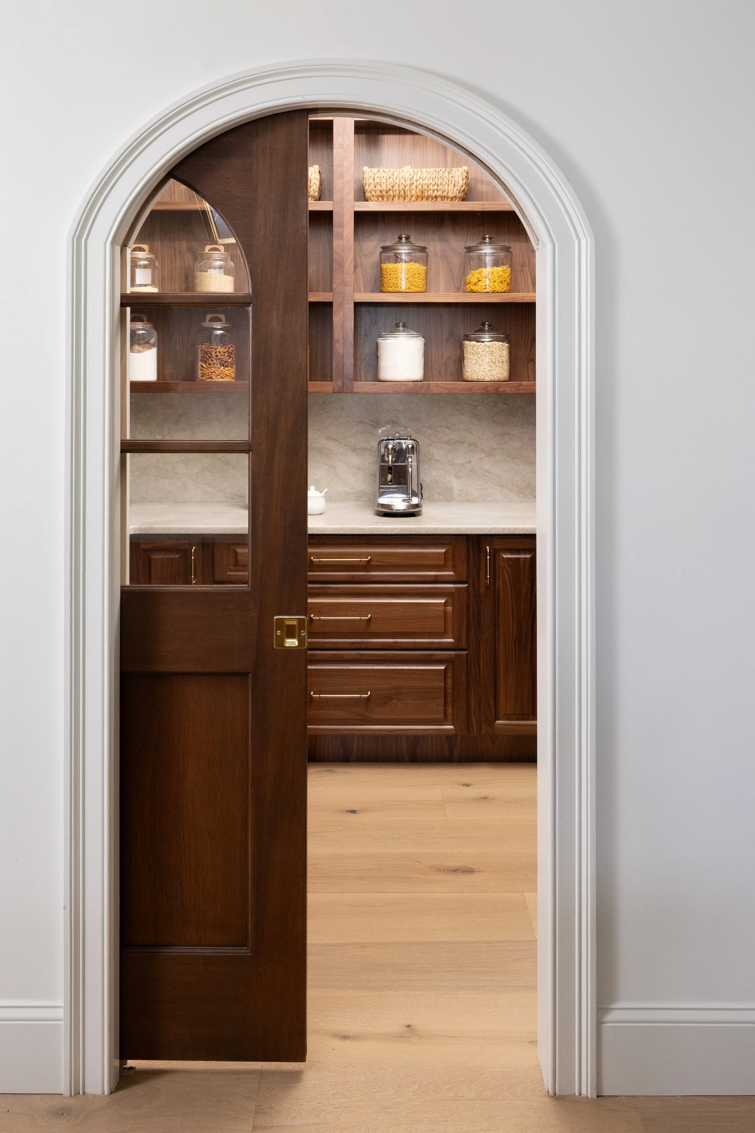 Open wooden door with glass panel revealing a kitchen with dark wood cabinetry, open shelves, and glass jars filled with pasta, cereal, and grains. Countertop with a coffee machine and a small teapot, with a white marbled backsplash.