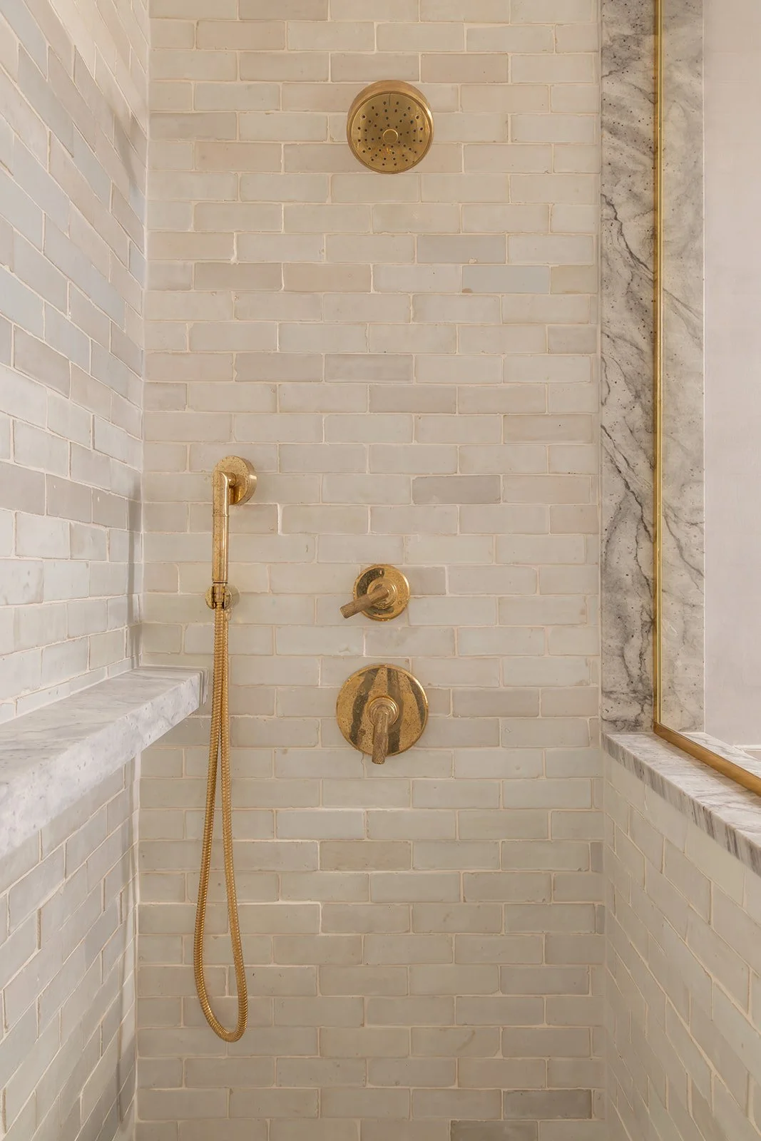 Inside view of a luxury stand-up shower showcasing light wall tiles, marble surfaces, a premium golden shower head, and a gold-framed glass door.
