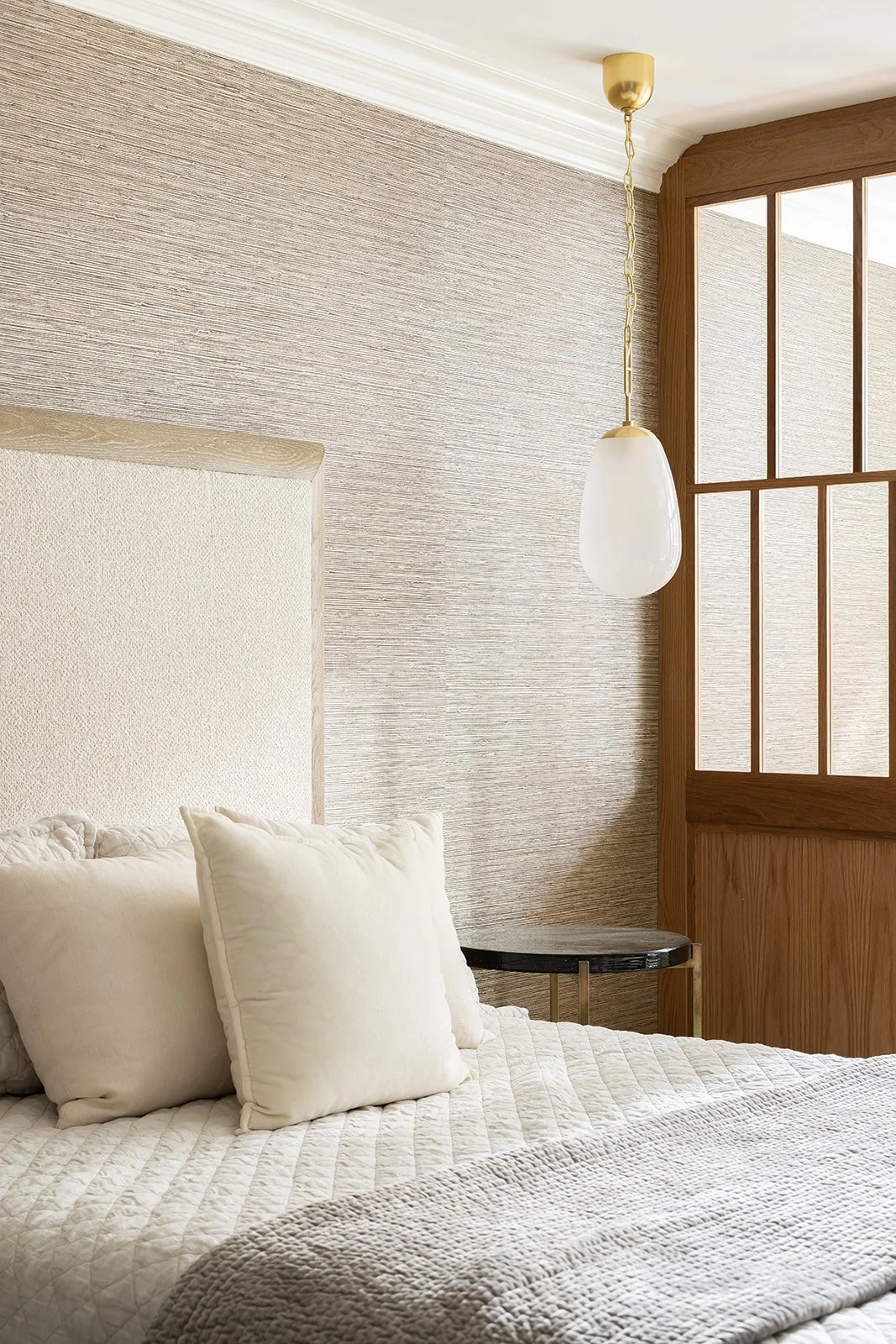 Modern bedroom corner styled with cream quilted bedding, a custom recessed headboard wall, textured dark beige wallpaper, a dark glass nightstand, and a hanging gold pendant light.