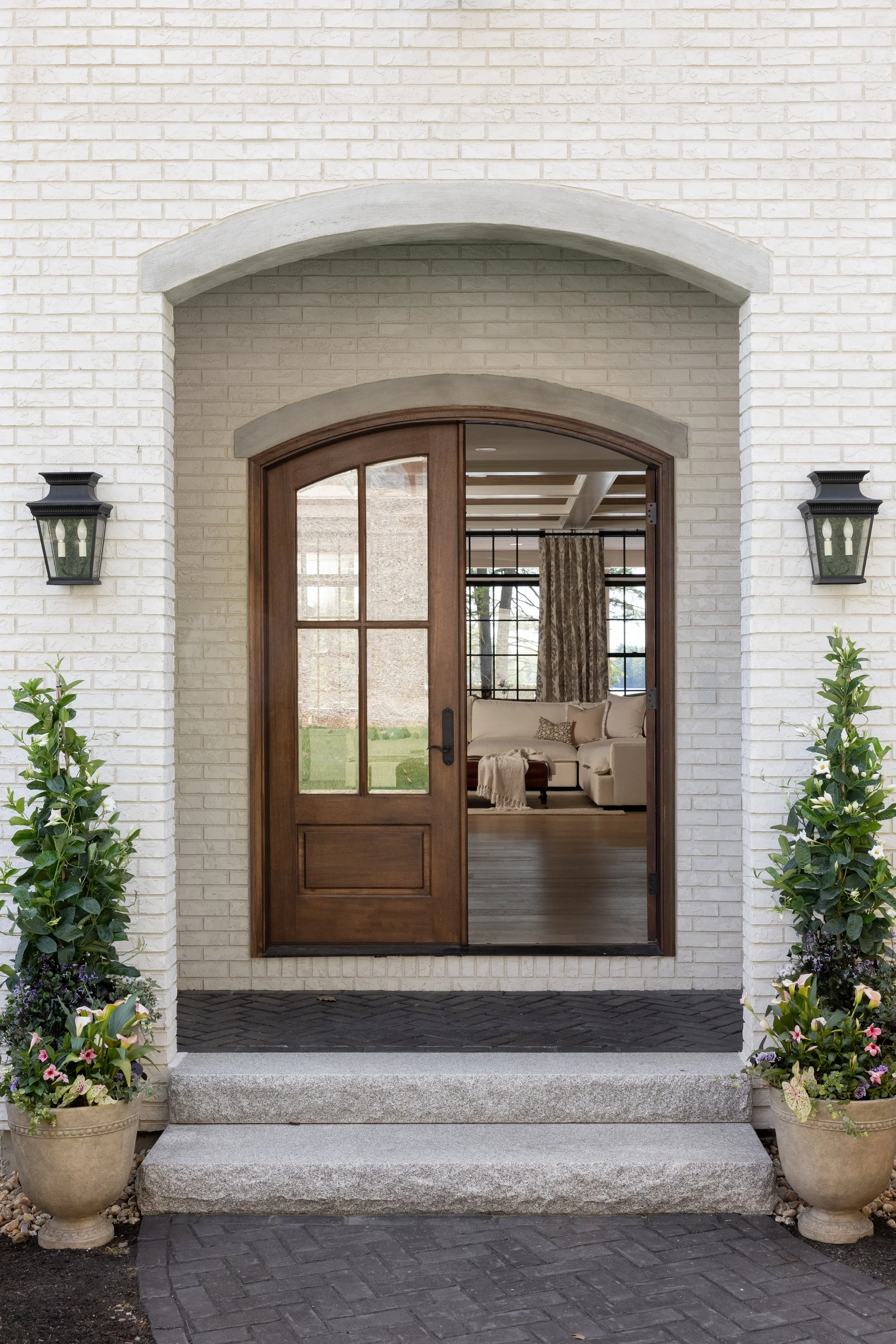 Front entryway of a house with a white brick exterior, a wooden door with glass panels, two black outdoor wall lanterns, potted plants on either side of the steps, and a view into a living room with beige furniture and windows.