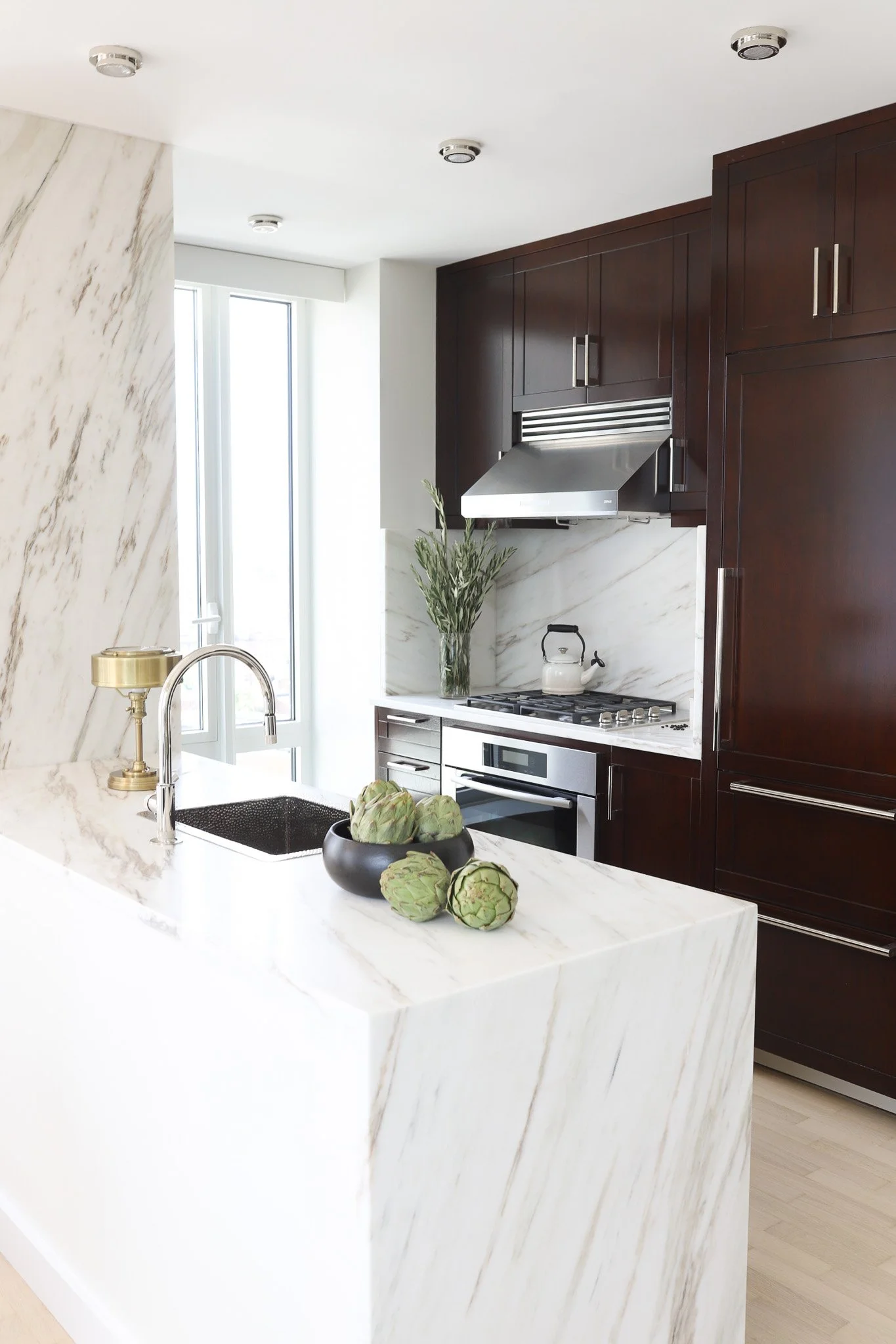 Bright luxury kitchen featuring Imperial Danby Honed Marble countertops, custom dark brown cabinets, matte silver appliances, and a gold lamp under natural window light.