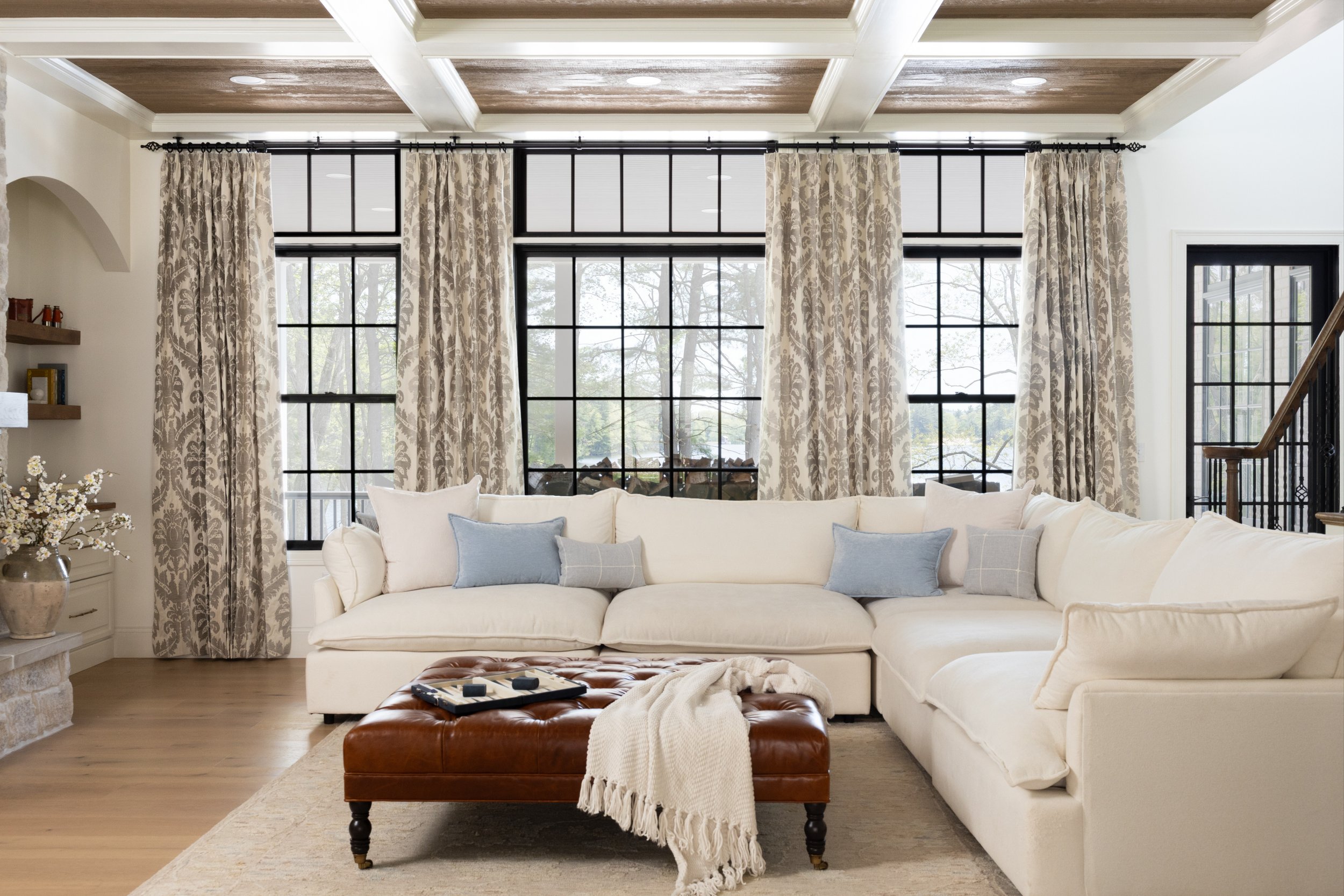 A living room with large windows, a white couch with light blue pillows, a brown leather ottoman with a cream throw blanket and a tray, a wooden shelf with books and decorations, and a ceiling with wood paneling and white beams.