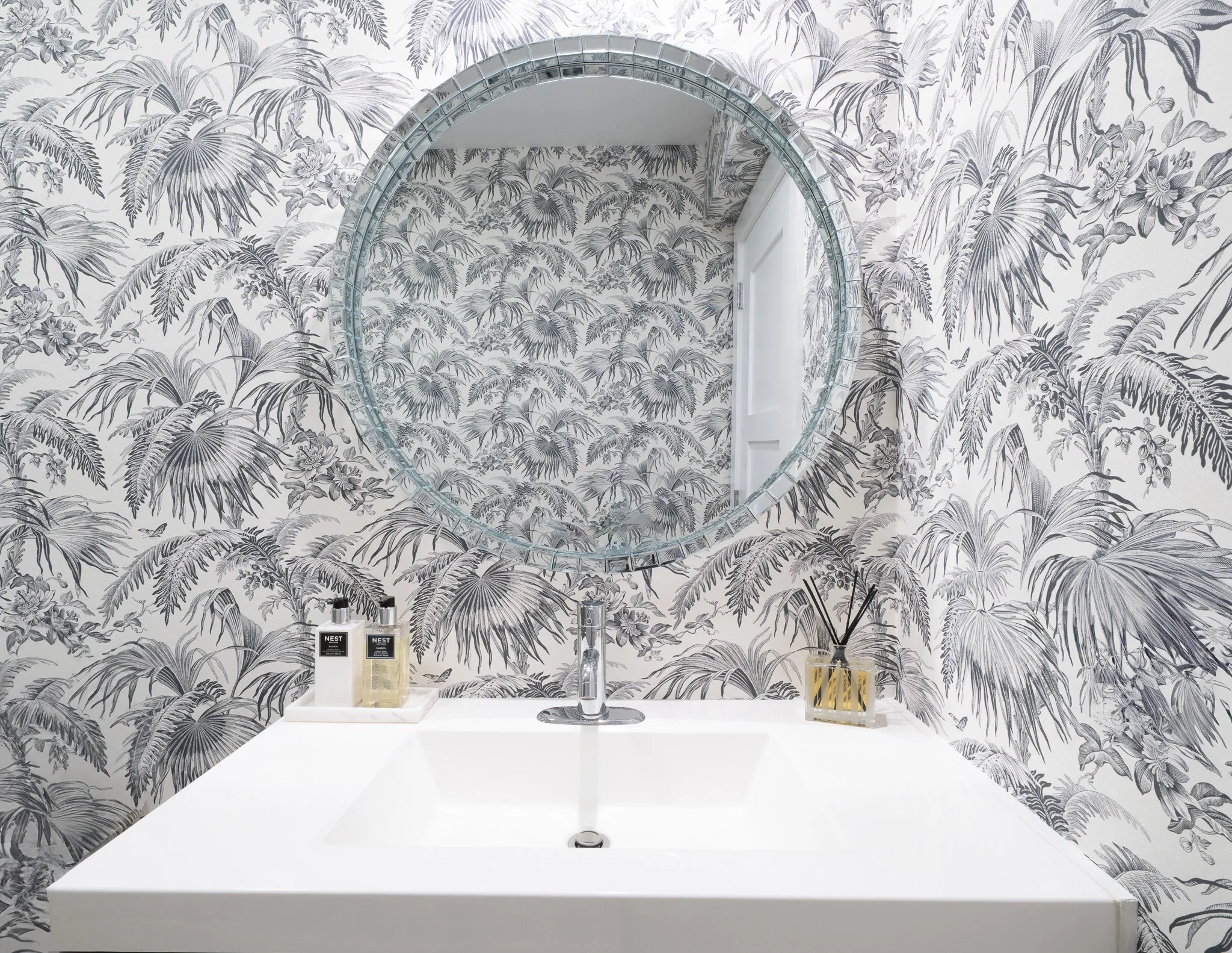 Modern bathroom vanity featuring a large round mirror, a white countertop, and dramatic black and white palm leaf patterned wallpaper.