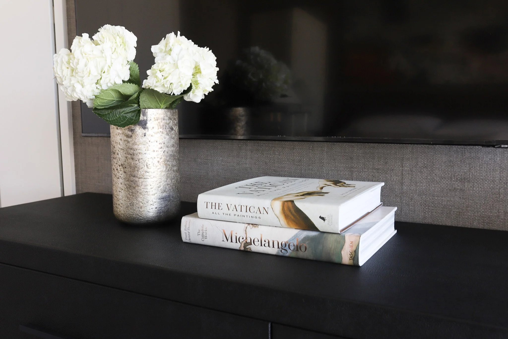 Close-up of a custom dark brown wood TV stand styled with a large vase of white flowers and stacked design books.