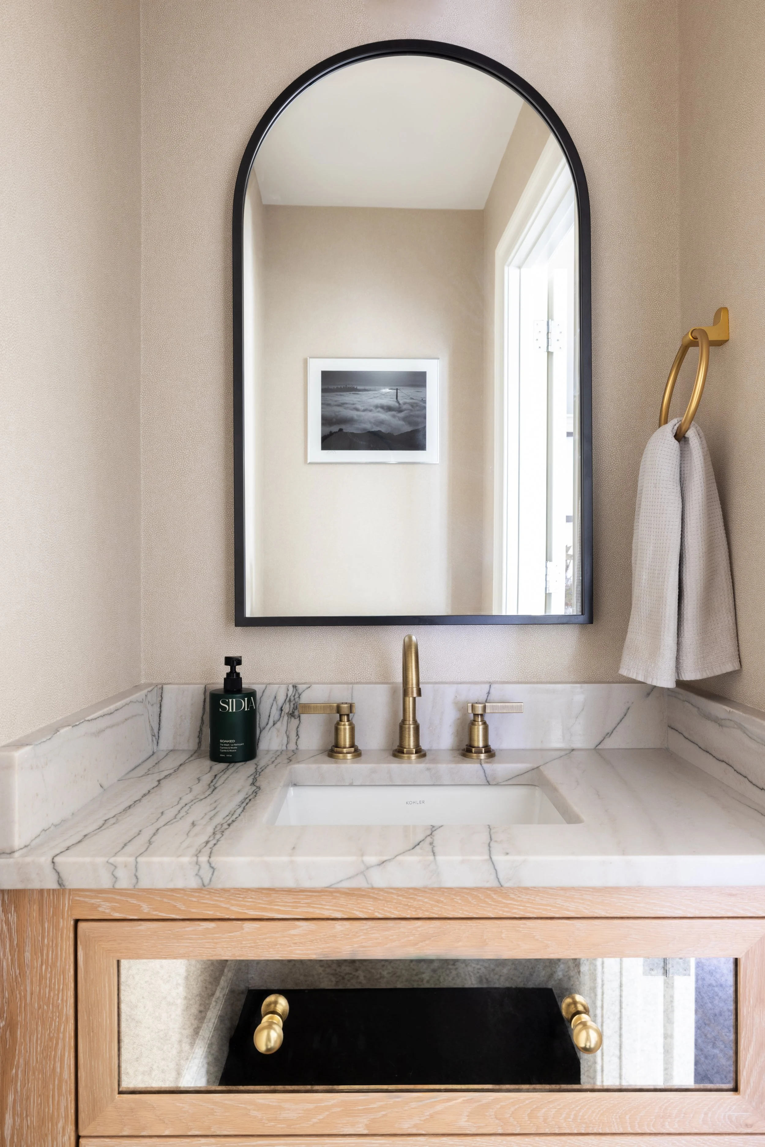  Elegant guest bathroom with a light marble sink and countertop, beige walls, a large mirror, and light brown cabinetry featuring dark glass drawers and gold knobs. 
