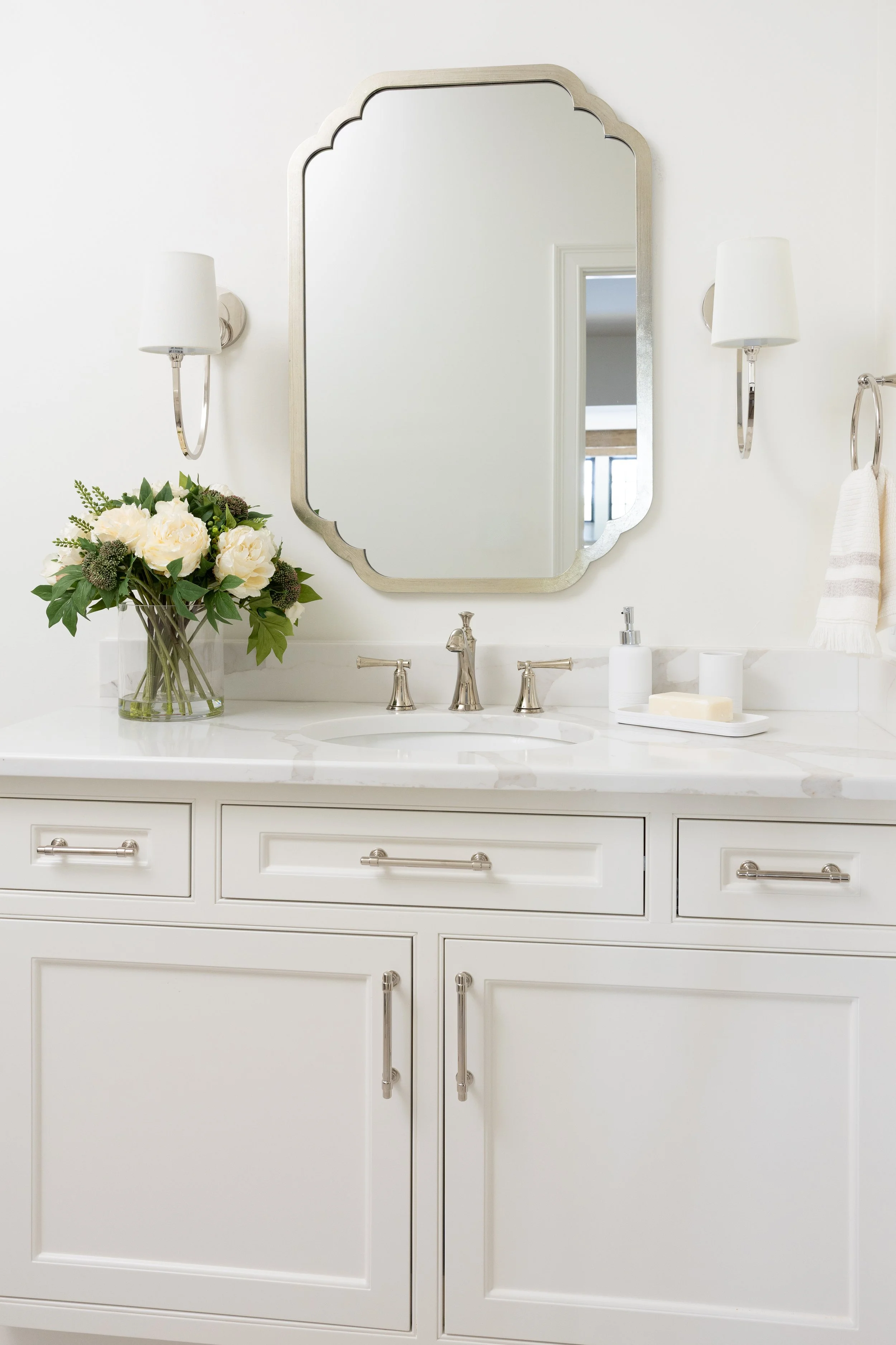 A modern white bathroom vanity with a marble countertop, a faucet, a vase with white flowers, a soap dispenser, a soap bar, and a mirror with wall sconces