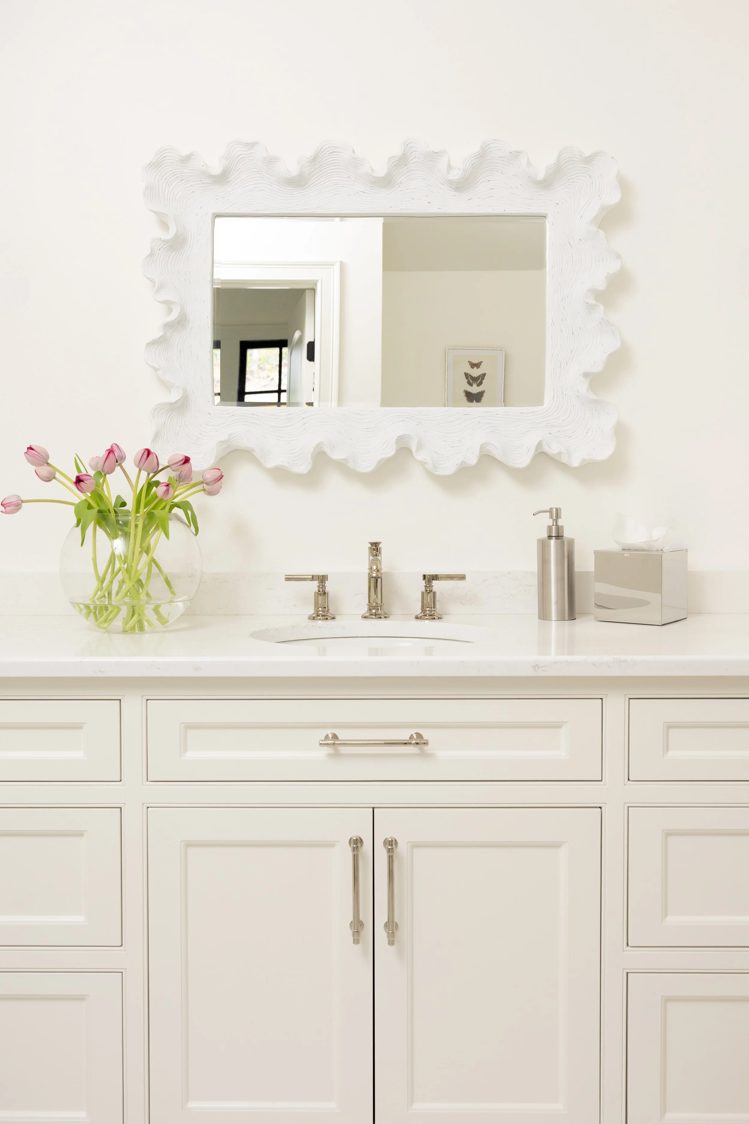Bathroom vanity with a white countertop, a vase of pink tulips, a mirror with a white textured frame, a tissue box, a soap dispenser, and a framed butterfly art on the wall.