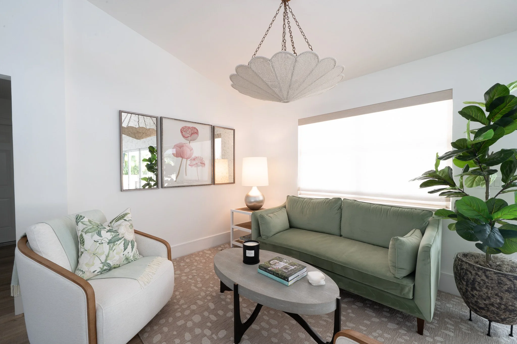 Elegant lounge area featuring a light green chenille sofa, white loveseat, a gray oval coffee table, a shell-shaped pendant light, and coastal art.