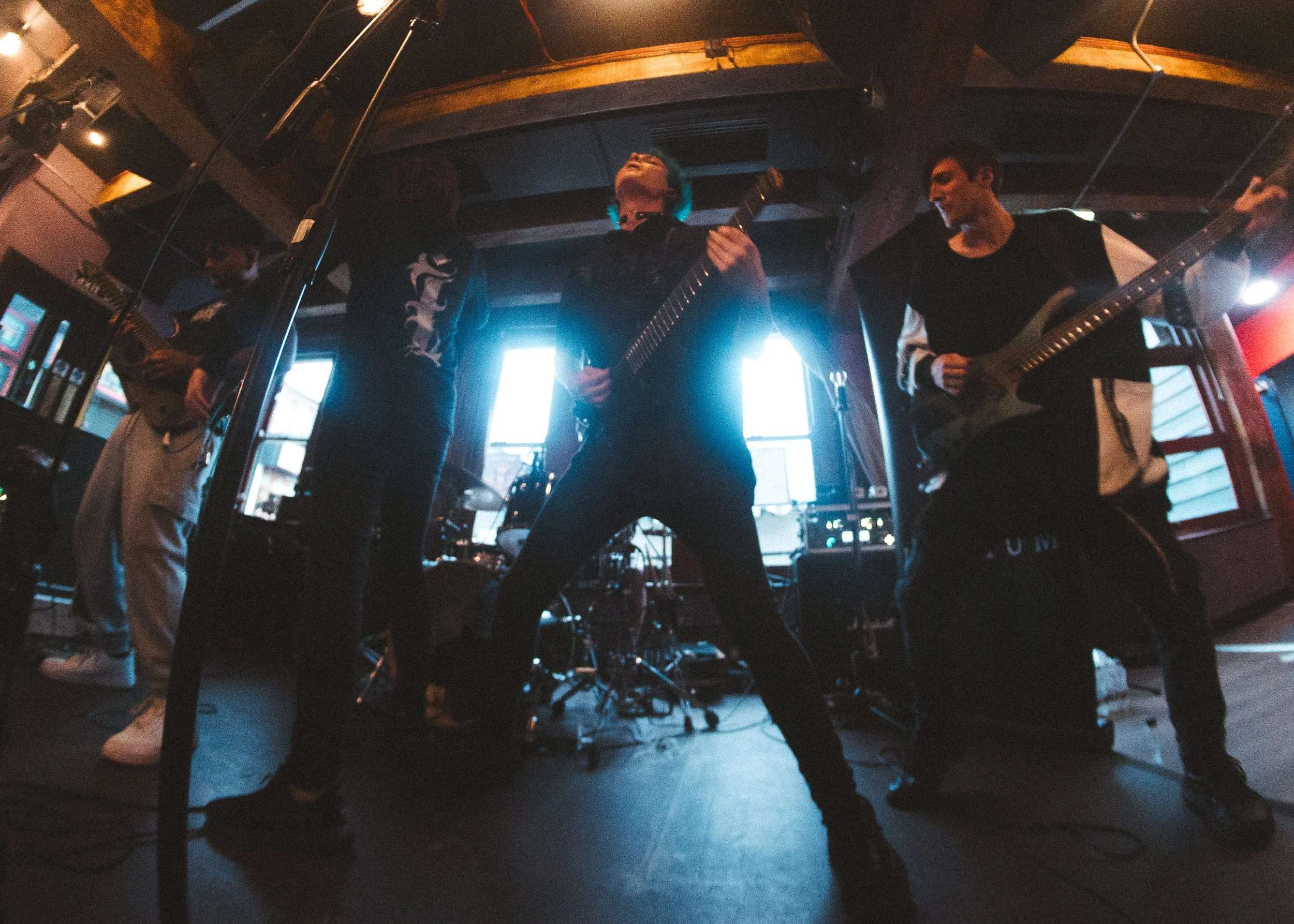 A band of four musicians performing on stage with guitars and drums in a dimly lit room, backlit by windows.