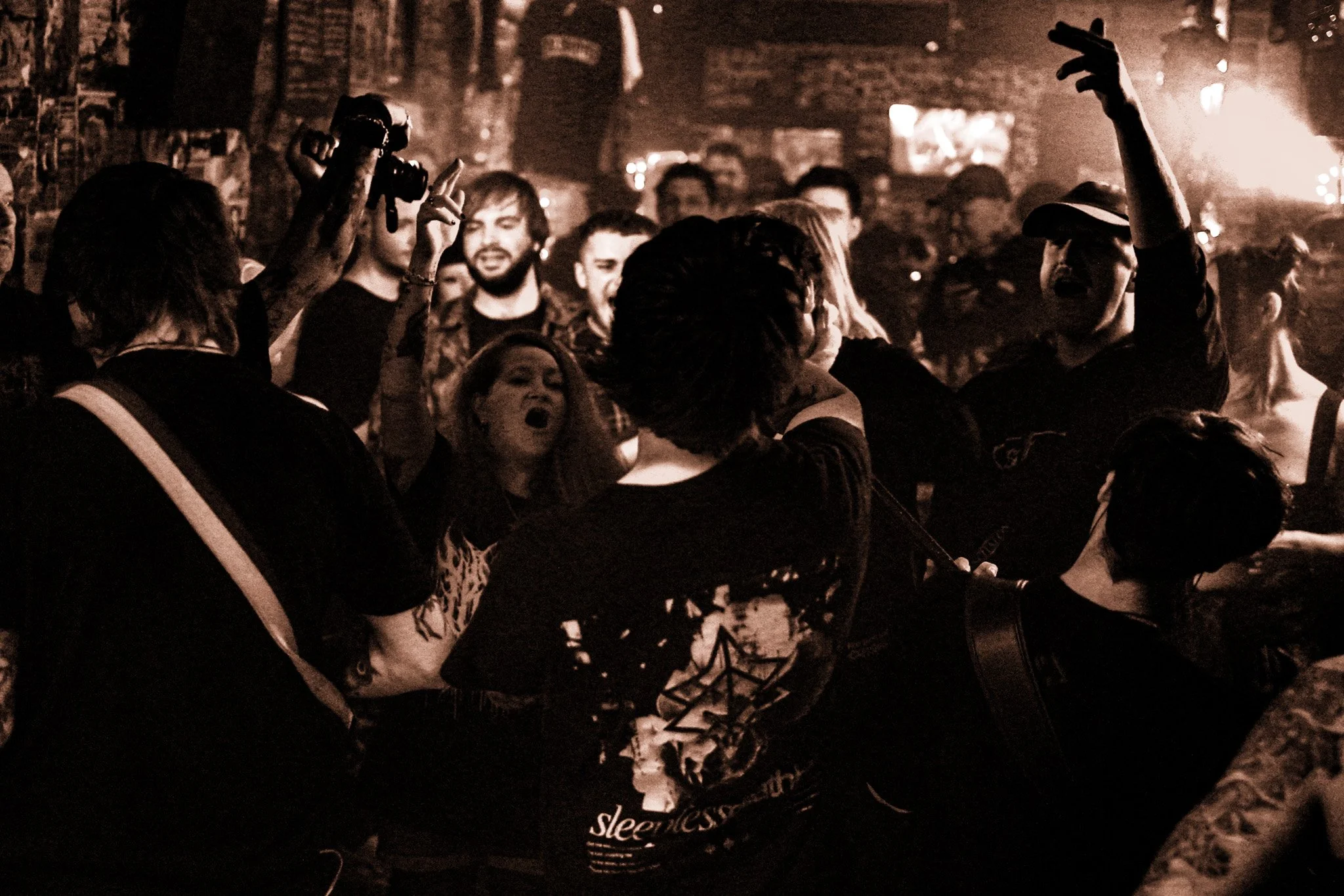 A crowd of people at a concert or music event, with some raising their hands and singing along. The scene is dark with warm lighting, and some individuals appear to be engaged in singing or shouting passionately.
