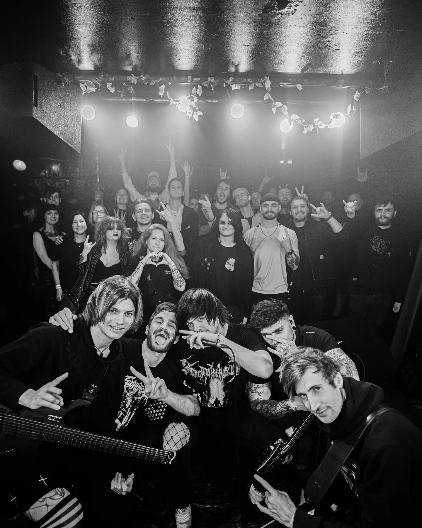 A black and white photo of a lively group of people at a concert or club, with some holding guitars, posing together and making peace signs or gestures, with a decorated ceiling and some stage lights in the background.