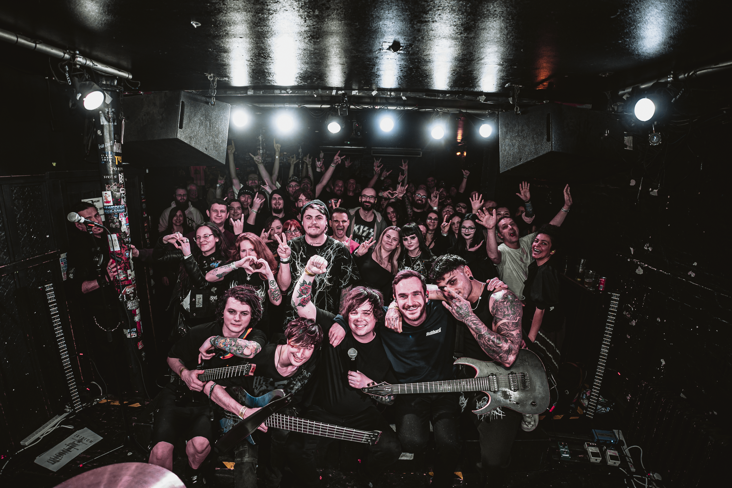 Group photo of a band with three guitarists and a singer holding a microphone, surrounded by cheering fans at a concert venue.