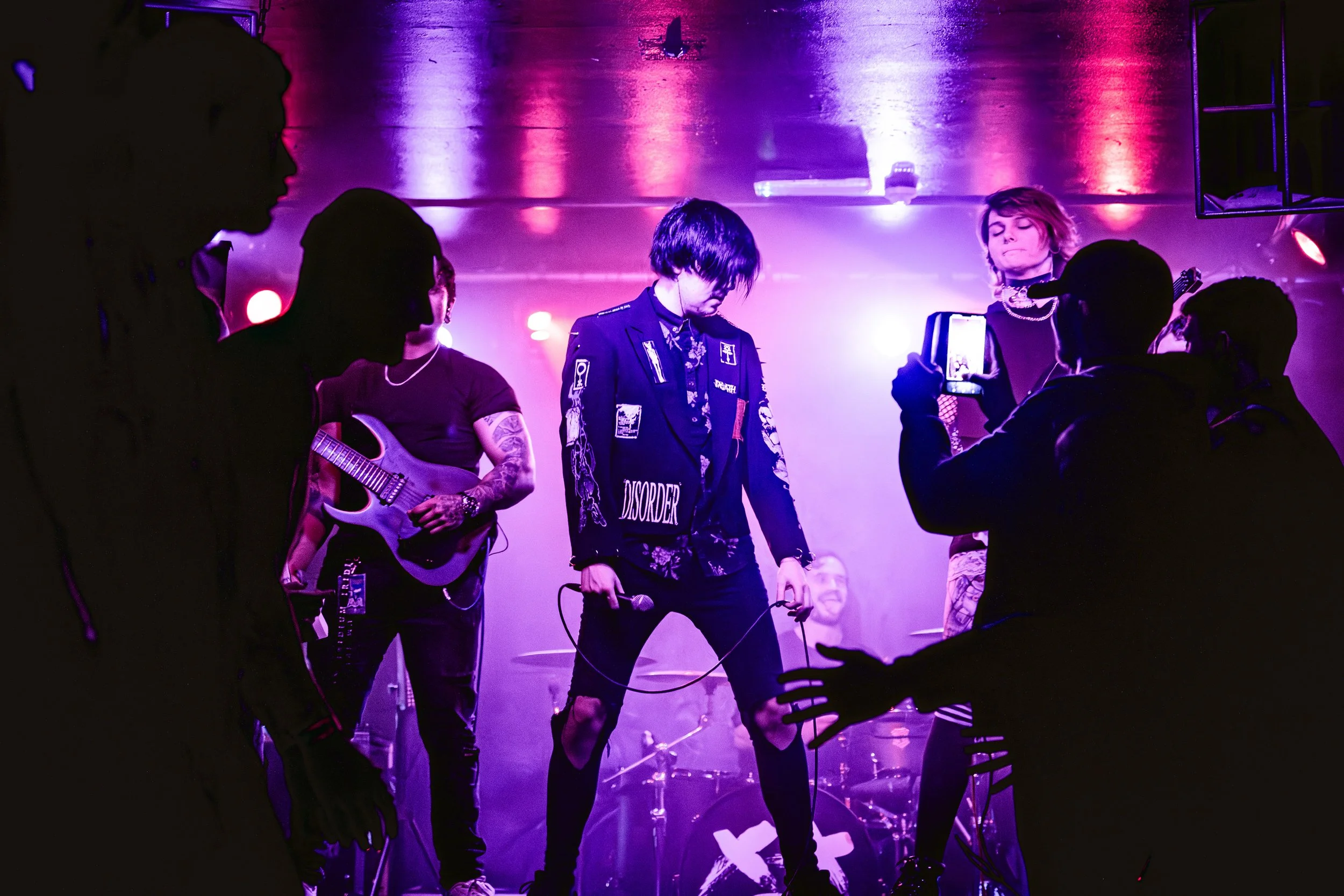 Music band performing on stage with purple and pink lighting, lead singer holding microphone, guitarist with tattoos, and a woman taking a photo.