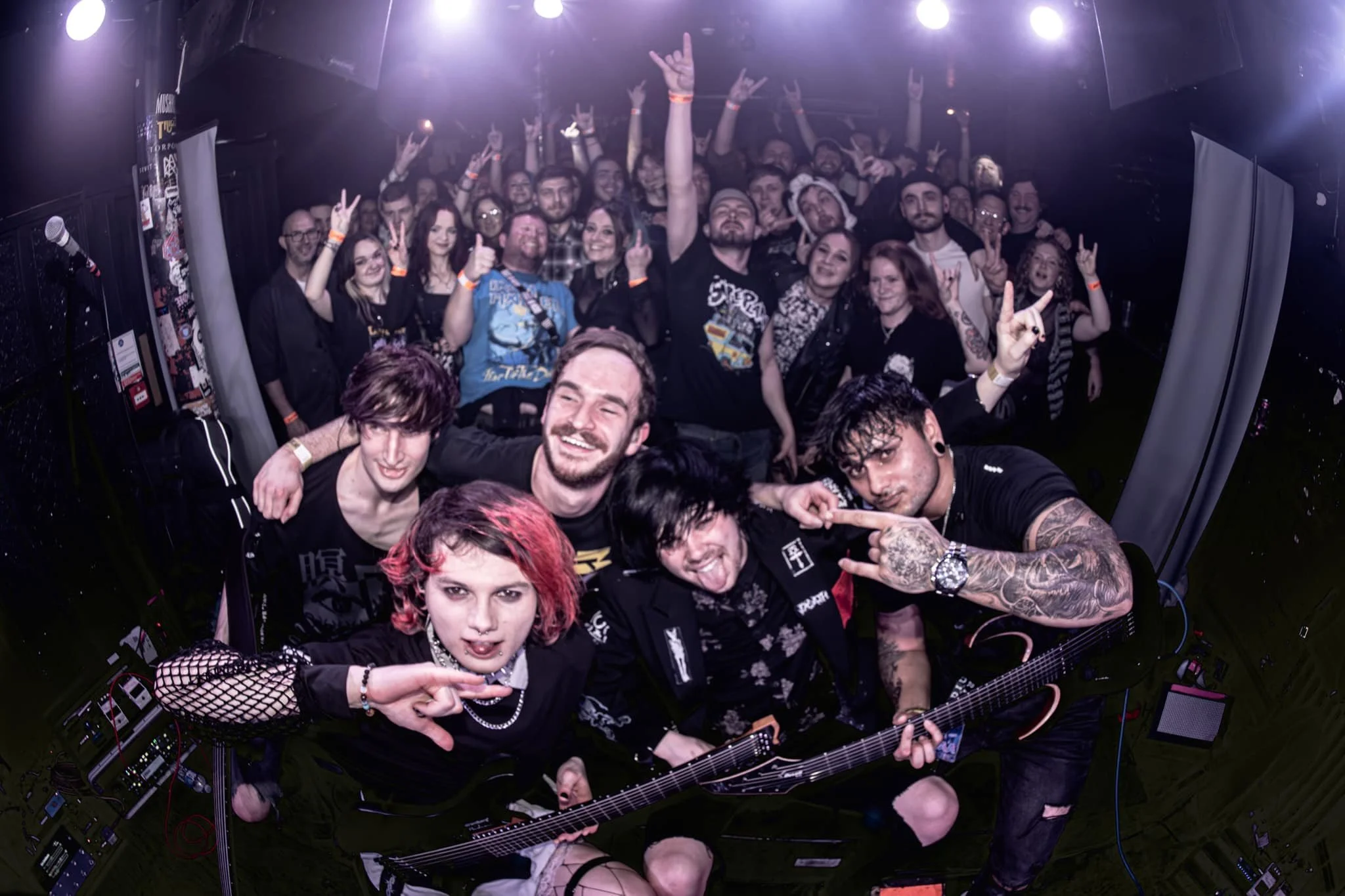Group of six musicians in the front of a crowd at a concert, with the audience making rock hand gestures and peace signs, smiling and enjoying the show.