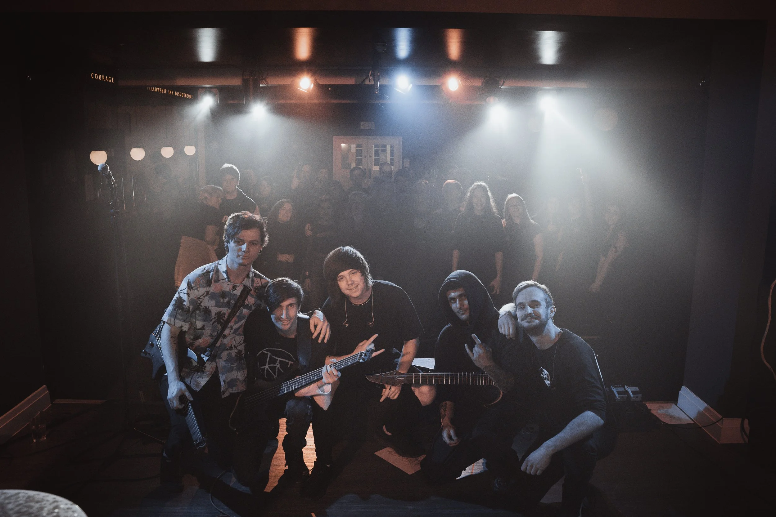 A group of five young men posing with musical instruments on a stage in front of an audience, with stage lights shining down on them and a crowd in the background.