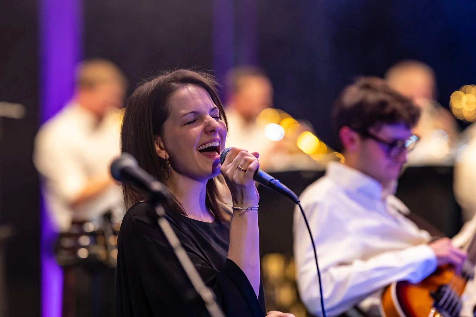 Eine junge Frau singt mit geschlossenen Augen an einem Mikrofon auf einer Bühne, neben einem Musiker mit E-Gitarre, bei einer Musikveranstaltung.