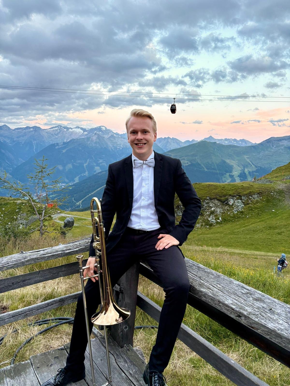 Ein junger Mann in formeller Kleidung mit Fliege sitzt auf einem Holzzaun in den Bergen und hält eine Posaune, mit einer bewölkten Berglandschaft im Hintergrund.