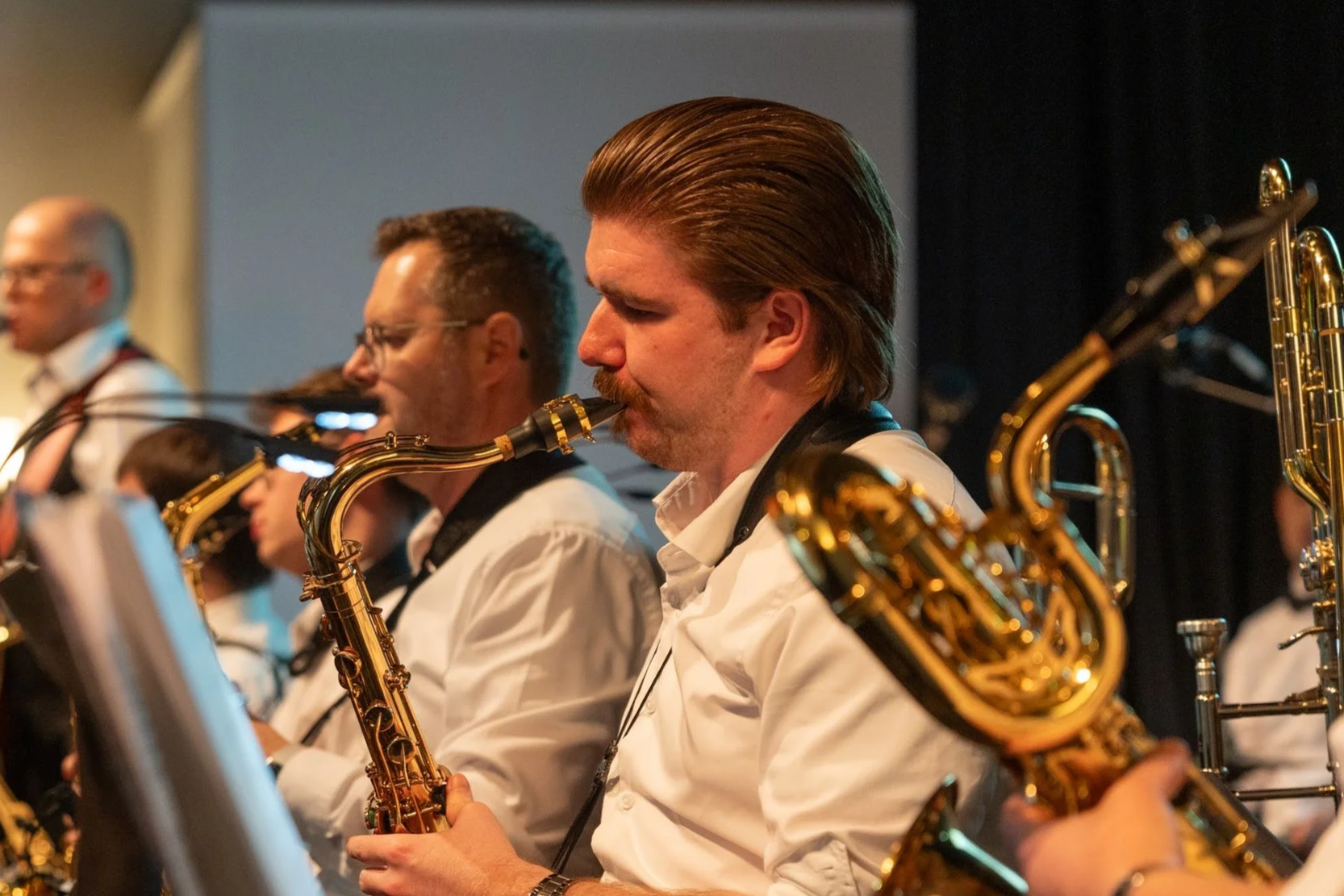 Musiker in einer Bigband spielen Saxophon, alle sind in weißen Hemden, im Hintergrund sind weitere Musiker zu sehen.