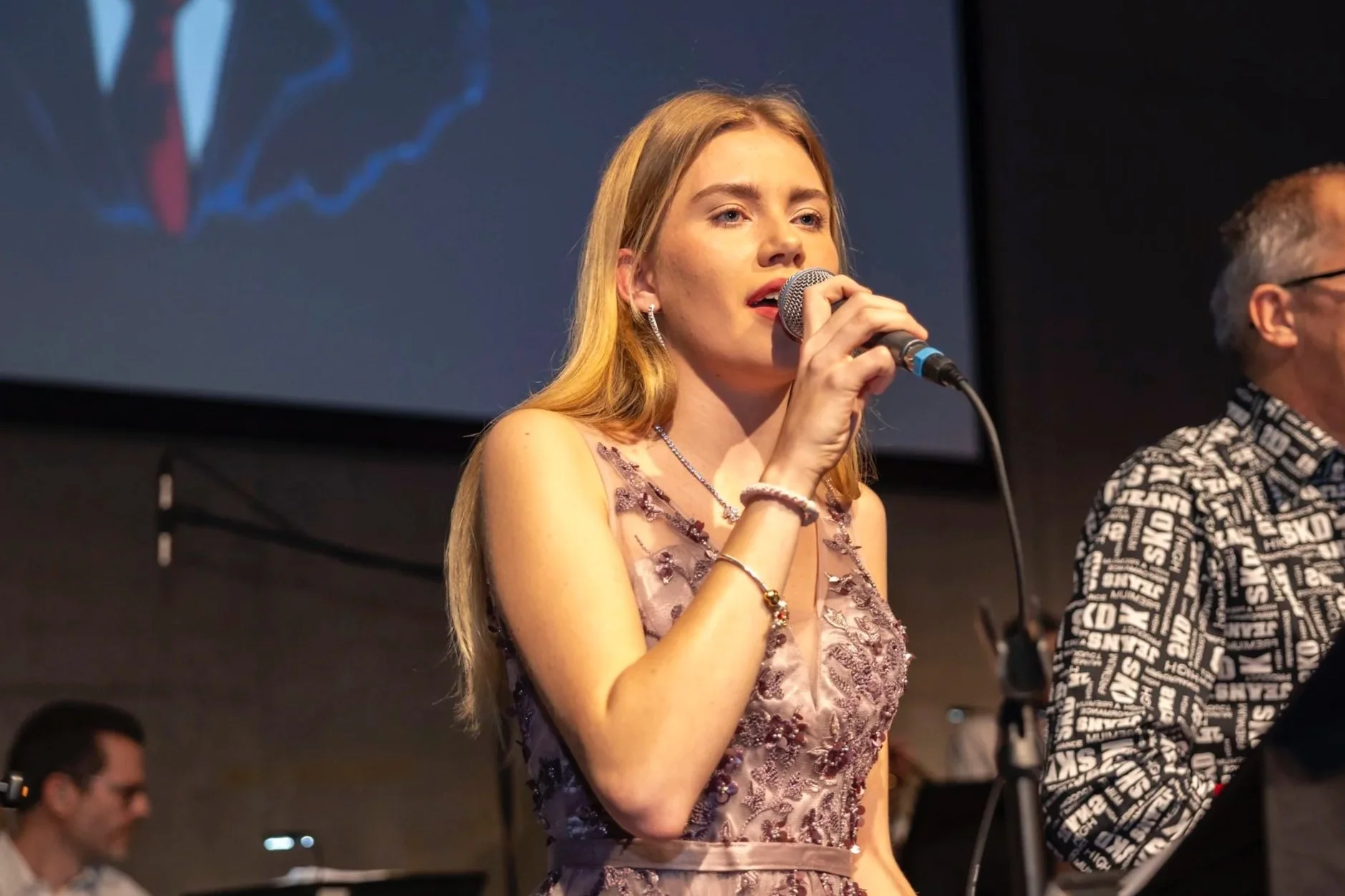 Junge Frau mit blonden Haaren singt mit Mikrofon auf einer Bühne, im Hintergrund ein Bildschirm und ein Mann mit Kopfhörer am Mischpult.