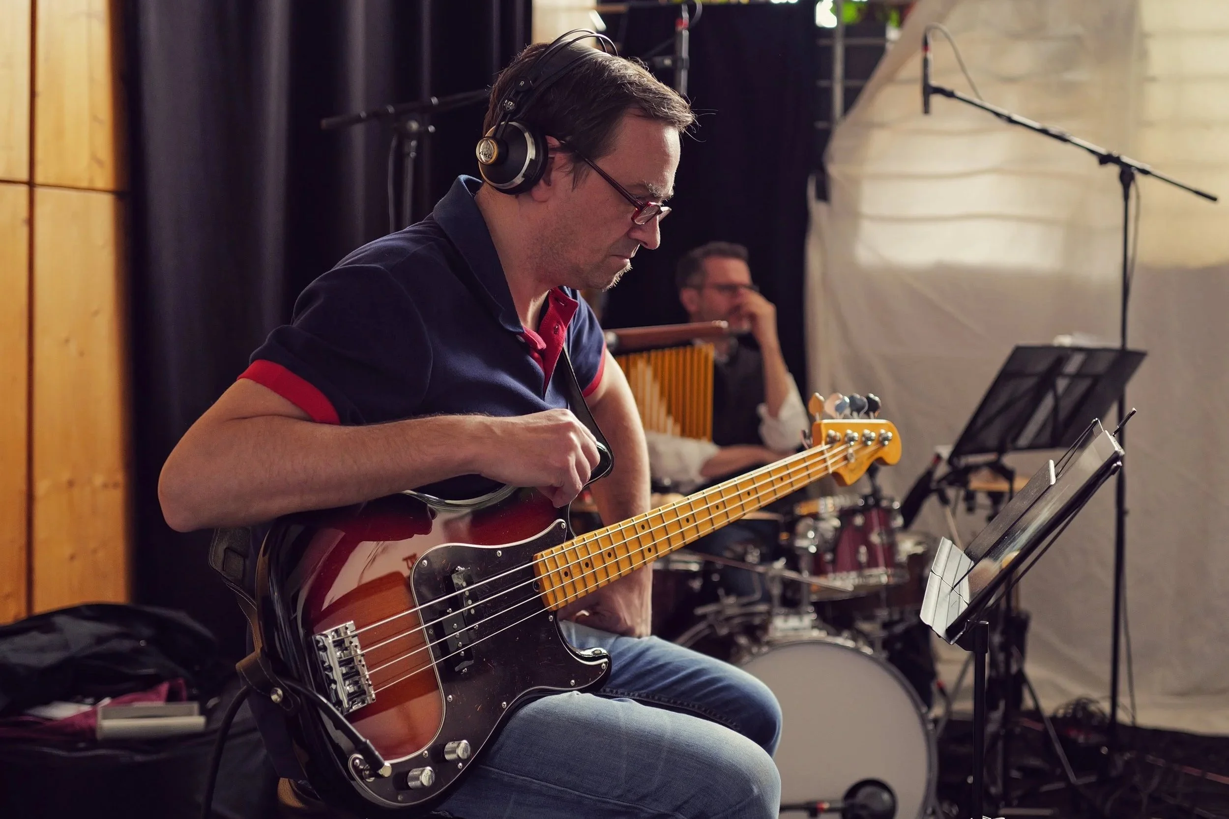 Ein Mann spielt Gitarre in einem Tonstudio, im Hintergrund steht ein Schlagzeuger. Beide tragen Kopfhörer und schauen auf Notenblätter.
