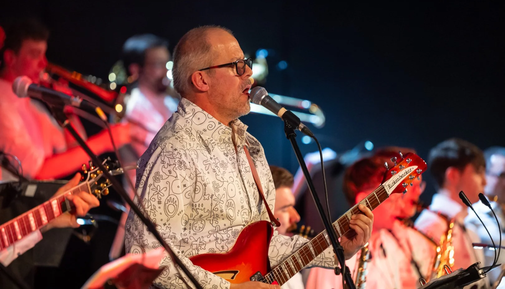 Musiker mit Brille und schwarzen Gitarrenspiel, gesessen in einem Orchester, singt in einem Konzertsaal, mit Bläsern im Hintergrund, auf einer Bühne mit schwarzem Vorhang und Musiktexten.