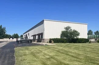 A large white industrial or commercial building with a small parking area in front, surrounded by a grassy lawn and a few trees under a clear blue sky.