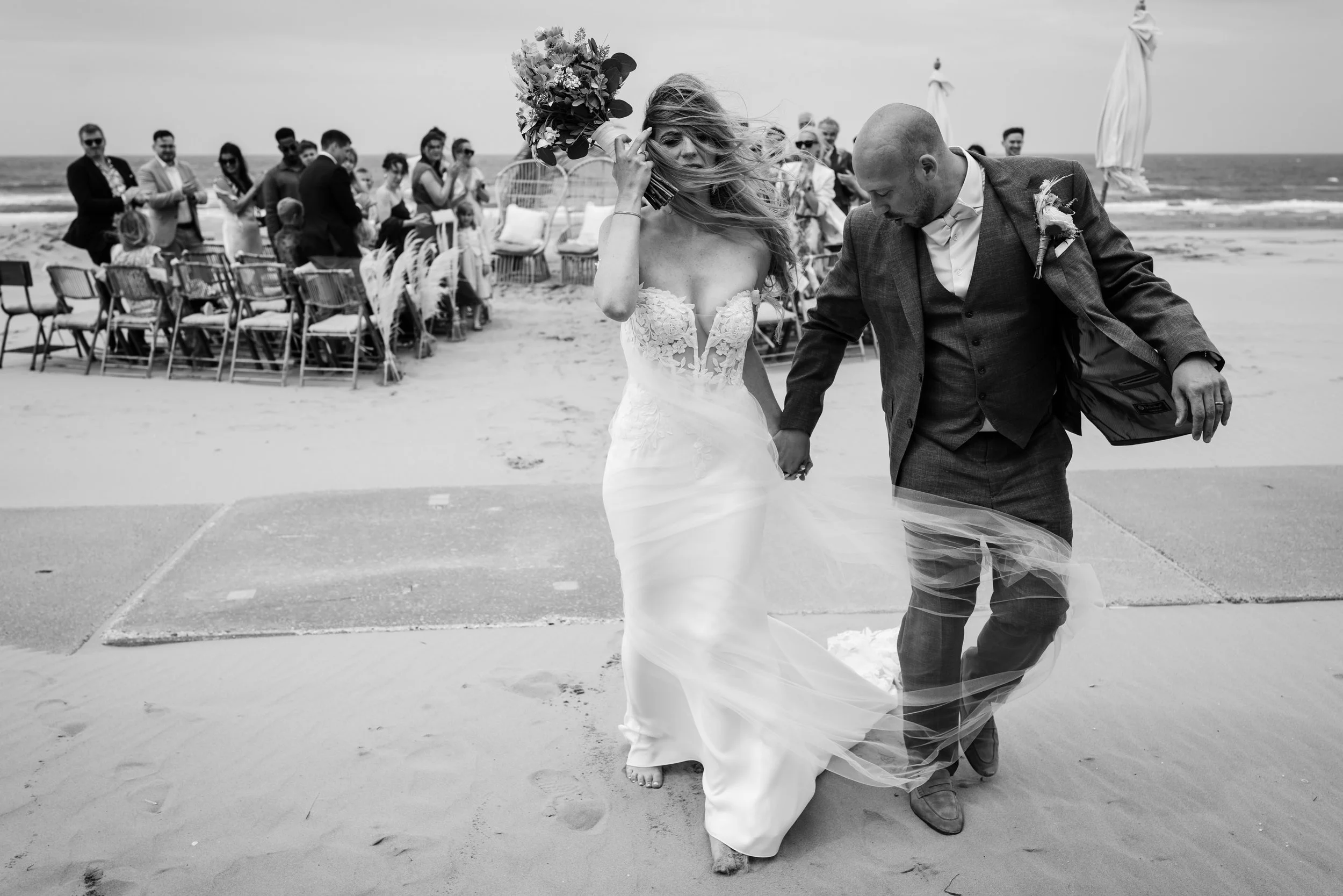 bruidspaar loopt van hun ceremonie op het strand weg, zwart wit, wind door het haar van de bruid