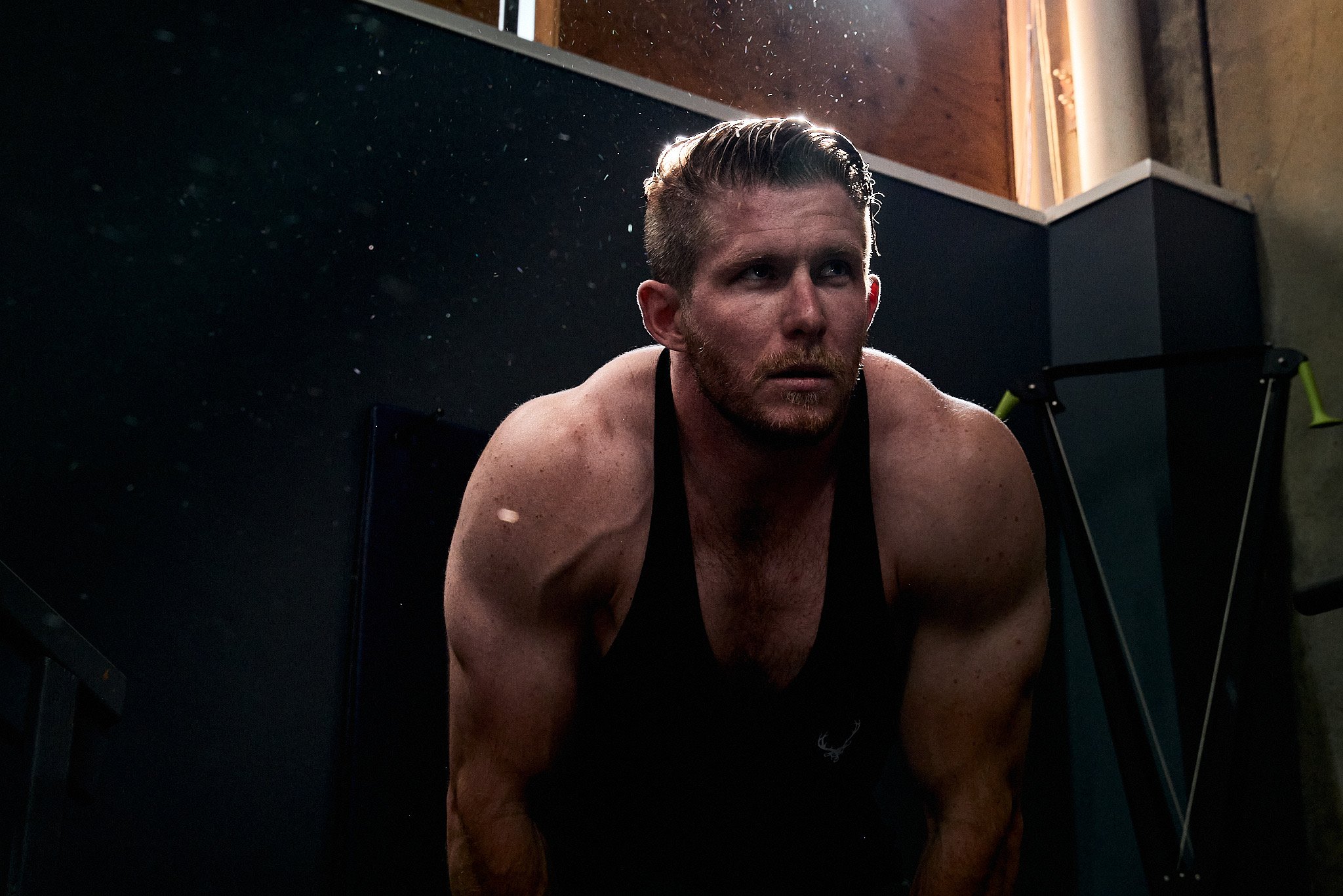 Portrait of a man in a black tank top, mid-workout at a gym. The man is standing in front of a dark background and is leaning over with a bright light shining above.