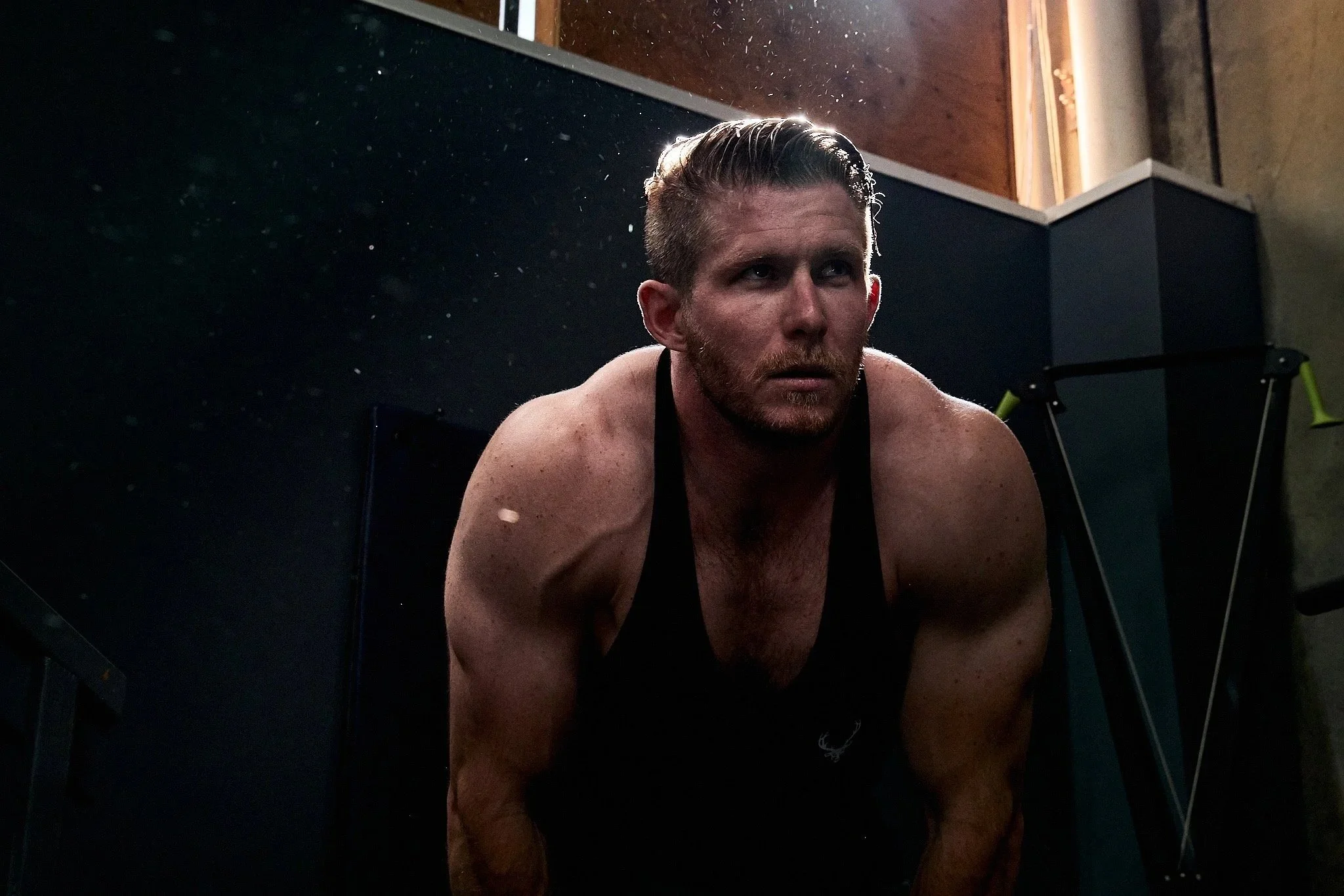 A man with wet hair and a beard, leaning forward, in a room with wooden and black walls, and gym equipment in the background.