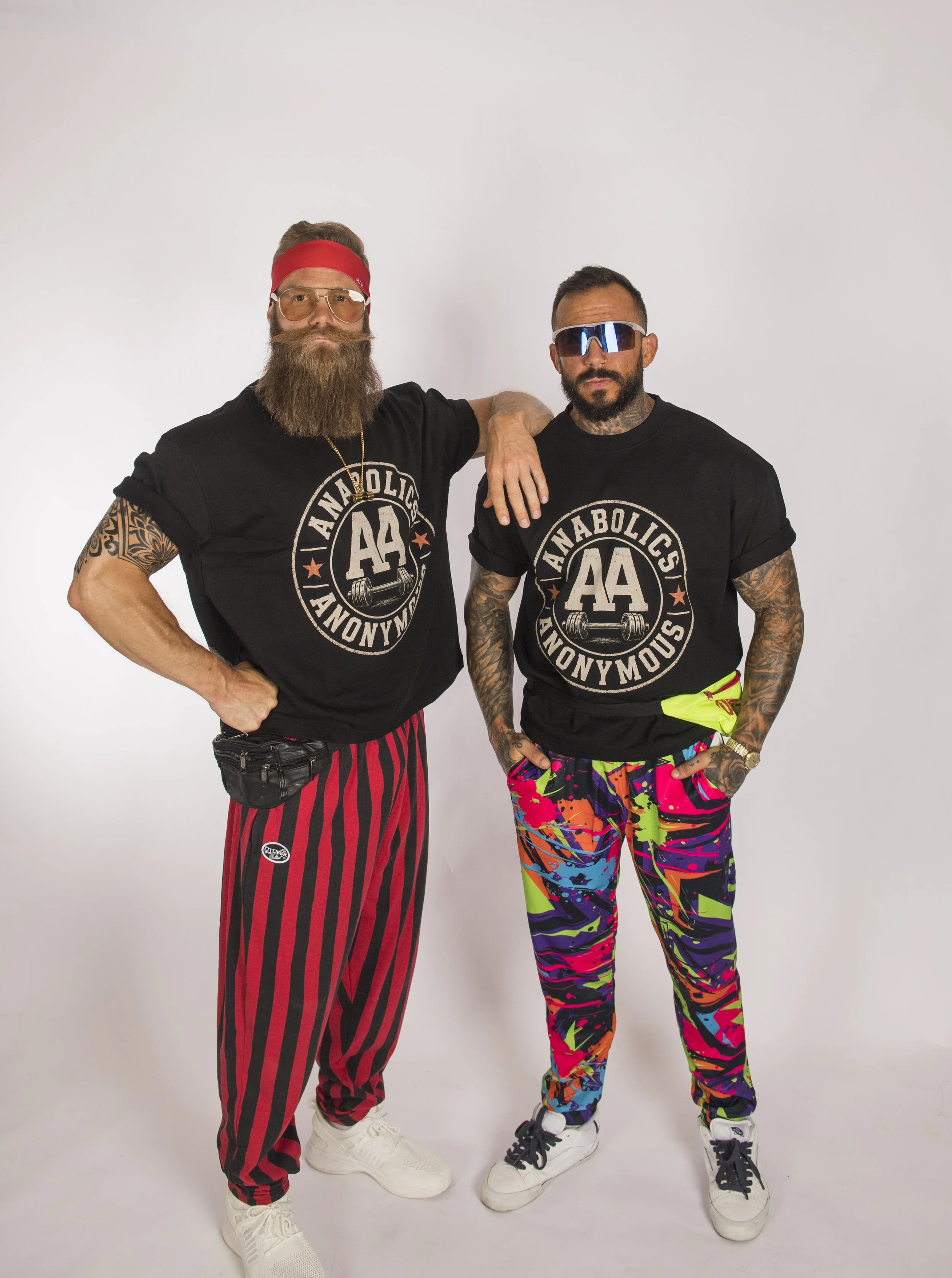 Two men standing against a white background wearing black T-shirts with the logo 'Anabolics Anonymous.' The man on the left has a full beard, glasses, a red headband, and red and black striped pants, while the man on the right has tattoos, sunglasses, colorful patterned pants, and assorted accessories.