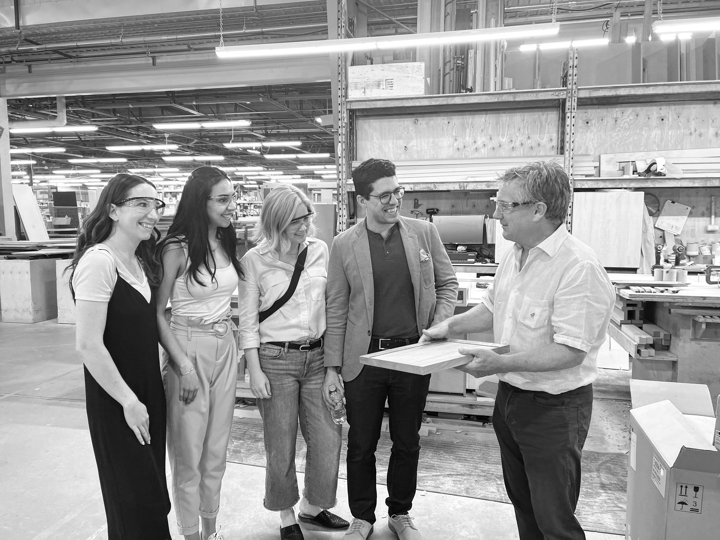Five people, four women and one man, in a workspace, smiling and looking at each other, with one man holding a wooden board. They are wearing safety glasses and casual clothing.