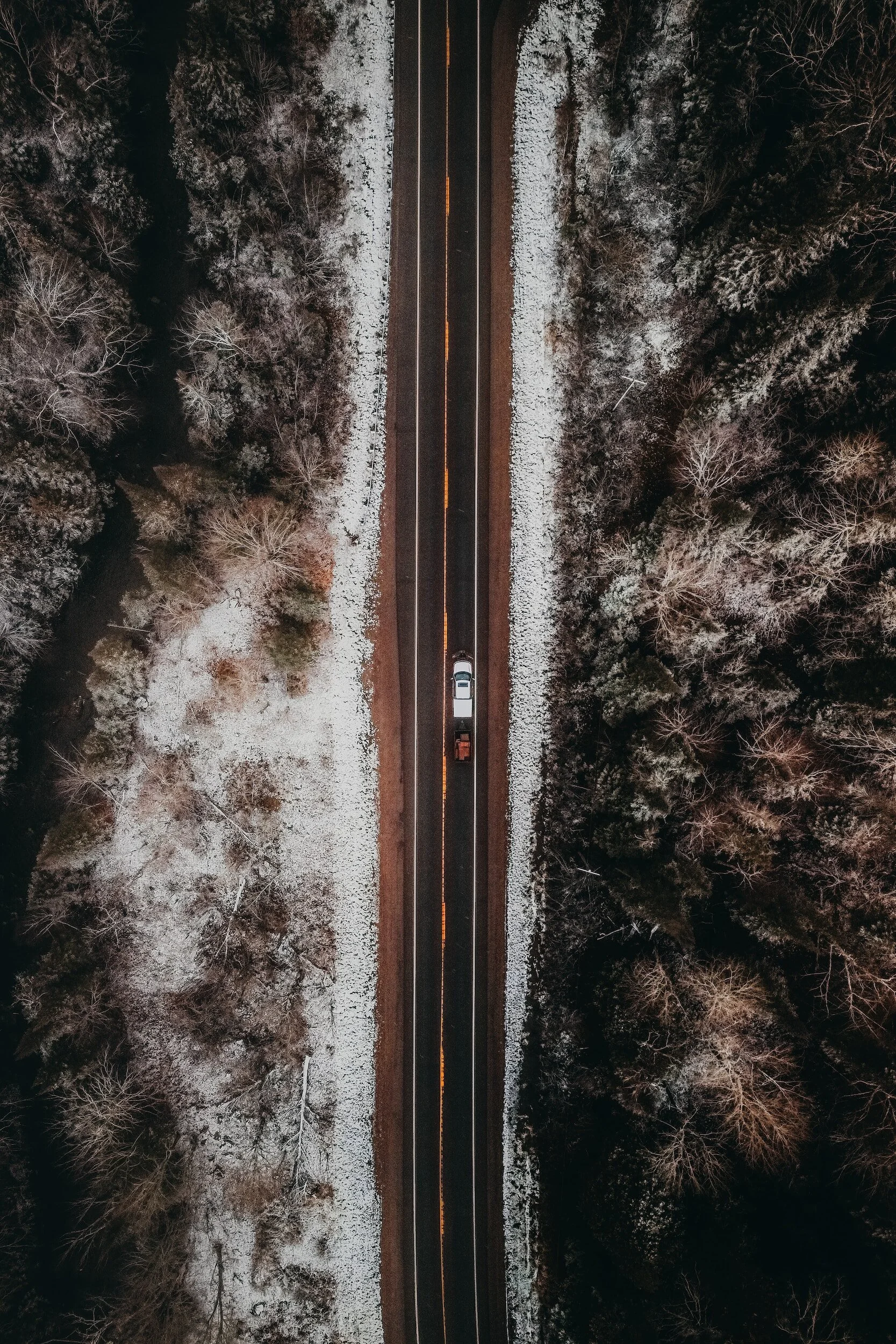 An aerial view of a straight road with a vehicle and a snow-covered forest on both sides during nighttime.