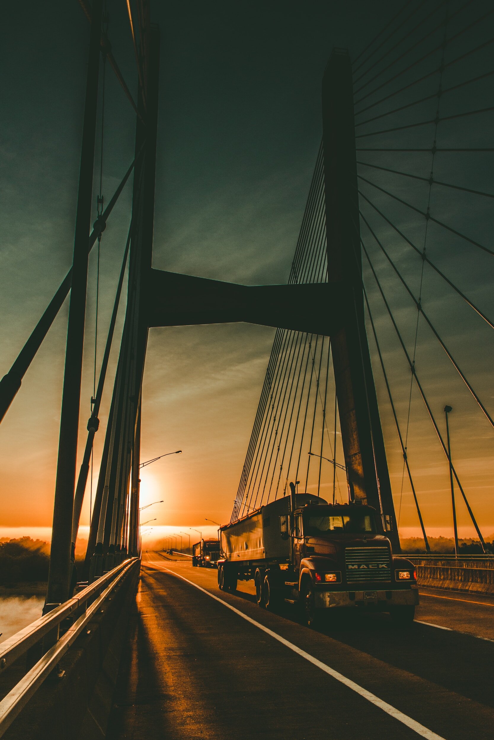A truck passing under a bridge on a highway at sunset, with streetlights lining the road.