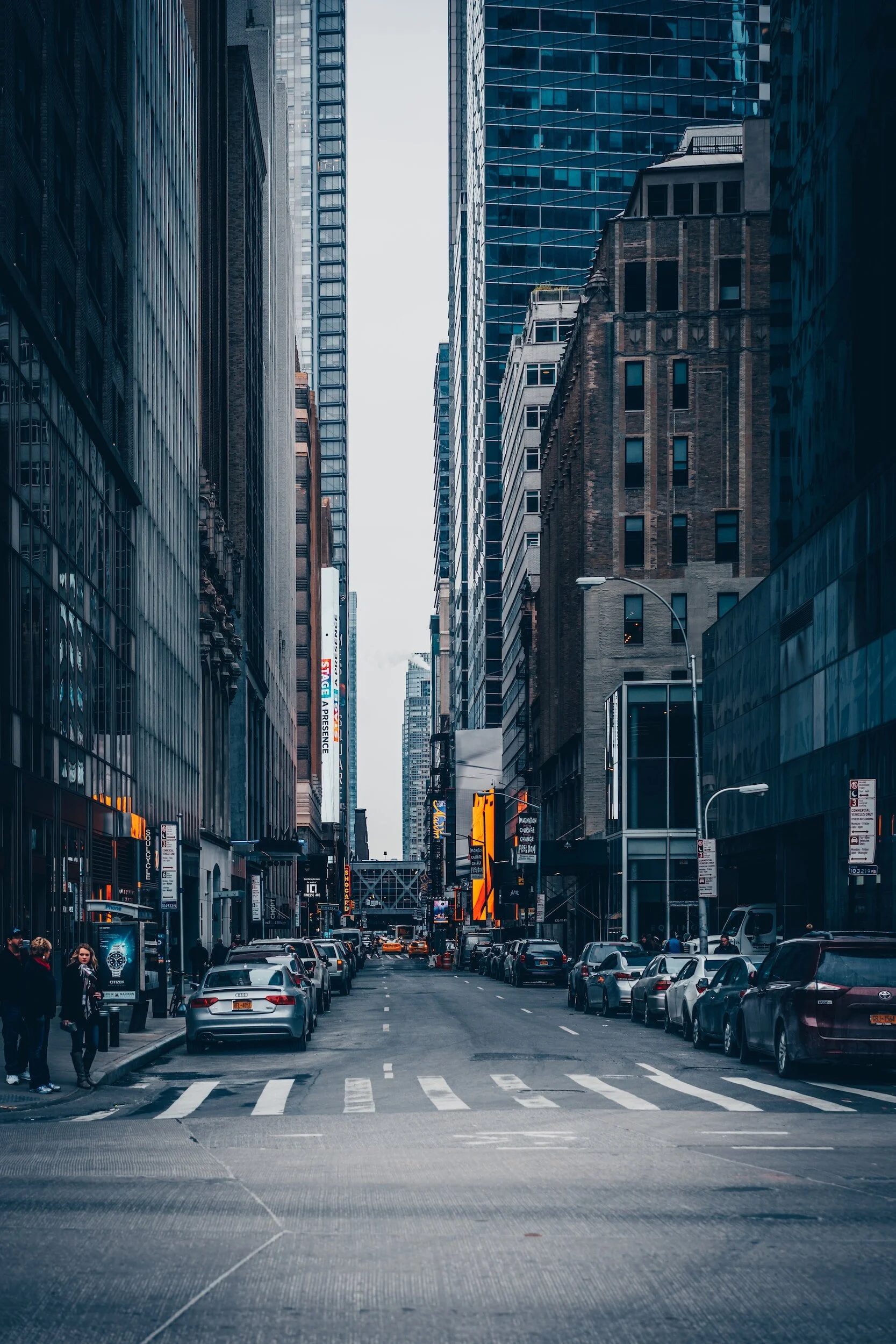 City street with tall buildings on both sides, parked cars along the curb, and a few pedestrians crossing the street in the foreground.